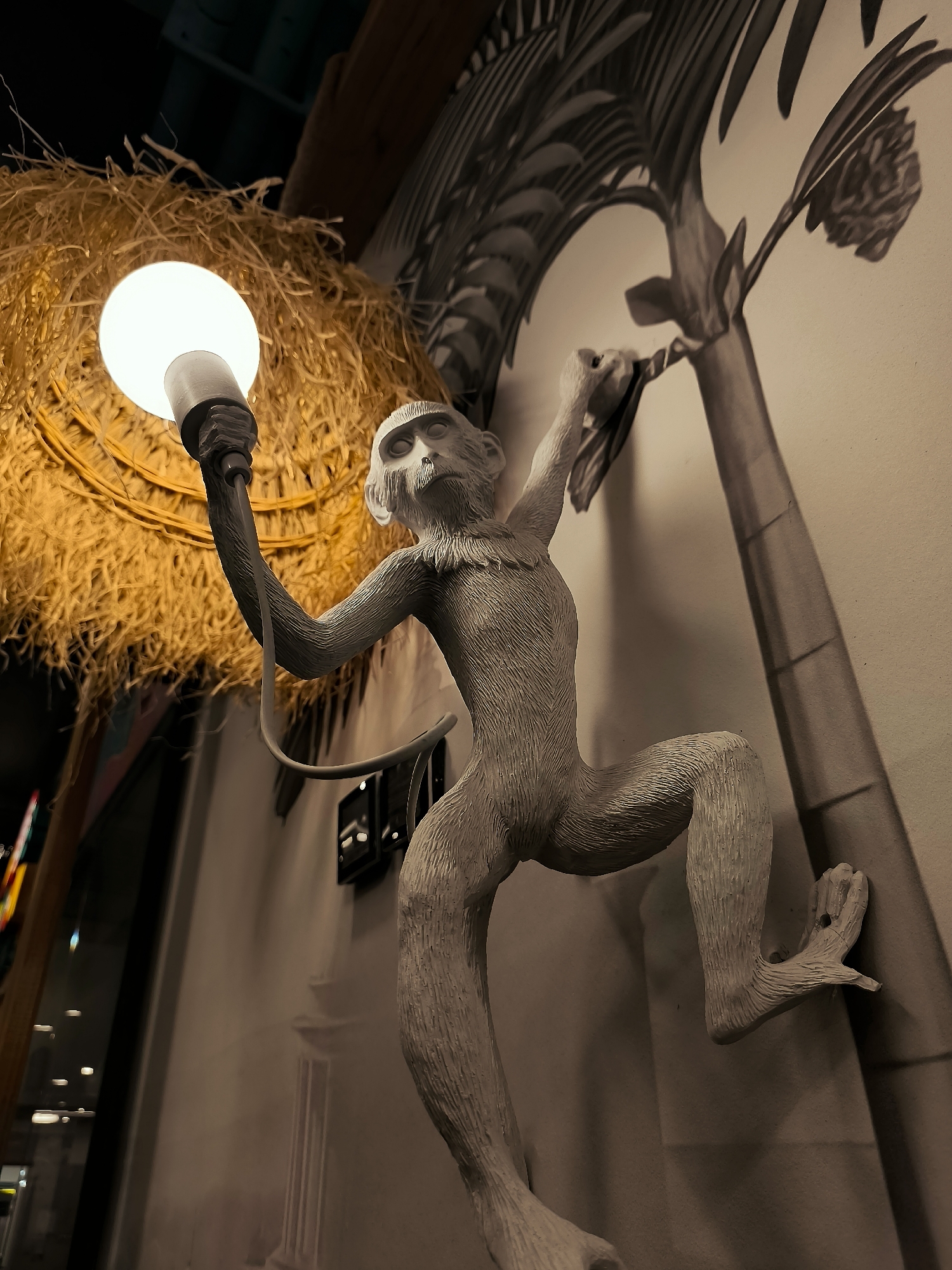 A realistic sculpture of a monkey is mounted on a wall. The monkey is depicted in a dynamic pose, with its arms outstretched, holding a glowing light bulb. It appears to be climbing or swinging, with one foot attached to the wall. Behind the monkey, a stylized mural of a palm tree can be seen. To the left, a nest-like structure made of straw surrounds another light fixture. The overall scene suggests an artistic installation, possibly in a restaurant or bar, with a jungle or tropical theme. The location is the city administration of Qarağandı city, Kazakhstan.