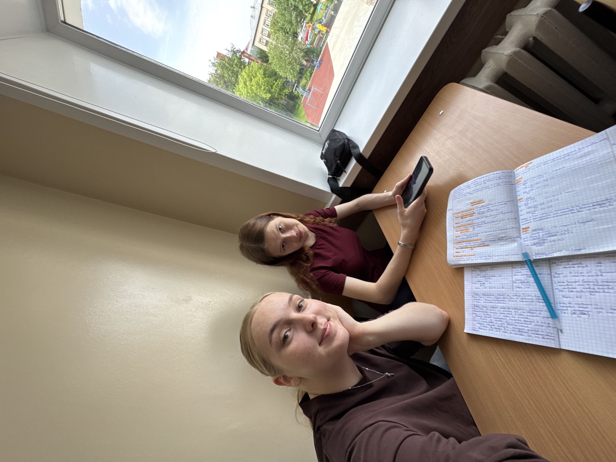 Two young women are seated at a desk near a window. The woman in the foreground is fair-skinned with blonde hair and wearing a brown long-sleeved shirt and silver chain. She is holding her chin and looking toward the camera. The woman in the background has reddish-brown hair and is wearing a maroon shirt. She is holding a black smartphone. On the desk are two notebooks with handwritten notes in Russian and a blue pen. Outside the window, a view of a residential area with trees and buildings is visible. Based on the style of the buildings and the vegetation, this appears to be in Perm, Russia. A black bag is seen on the window sill. A radiator is visible behind the desk.