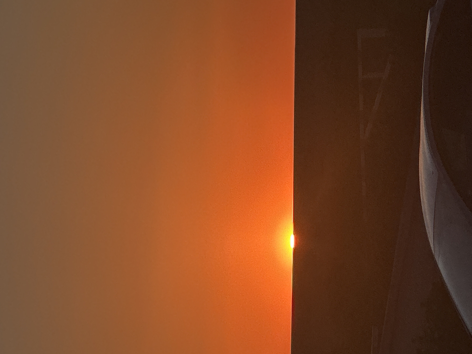 The image shows a sunrise. The sky is a gradient of orange to brown. On the right, a dark object with what appears to be a ladder or structural support and the curved edge of an object are silhouetted against the sky. There is a bright point of light, likely the sun, peeking between the vertical dark edge and the orange sky. The exact location within Khabarovsk, Russia, cannot be determined from the image.