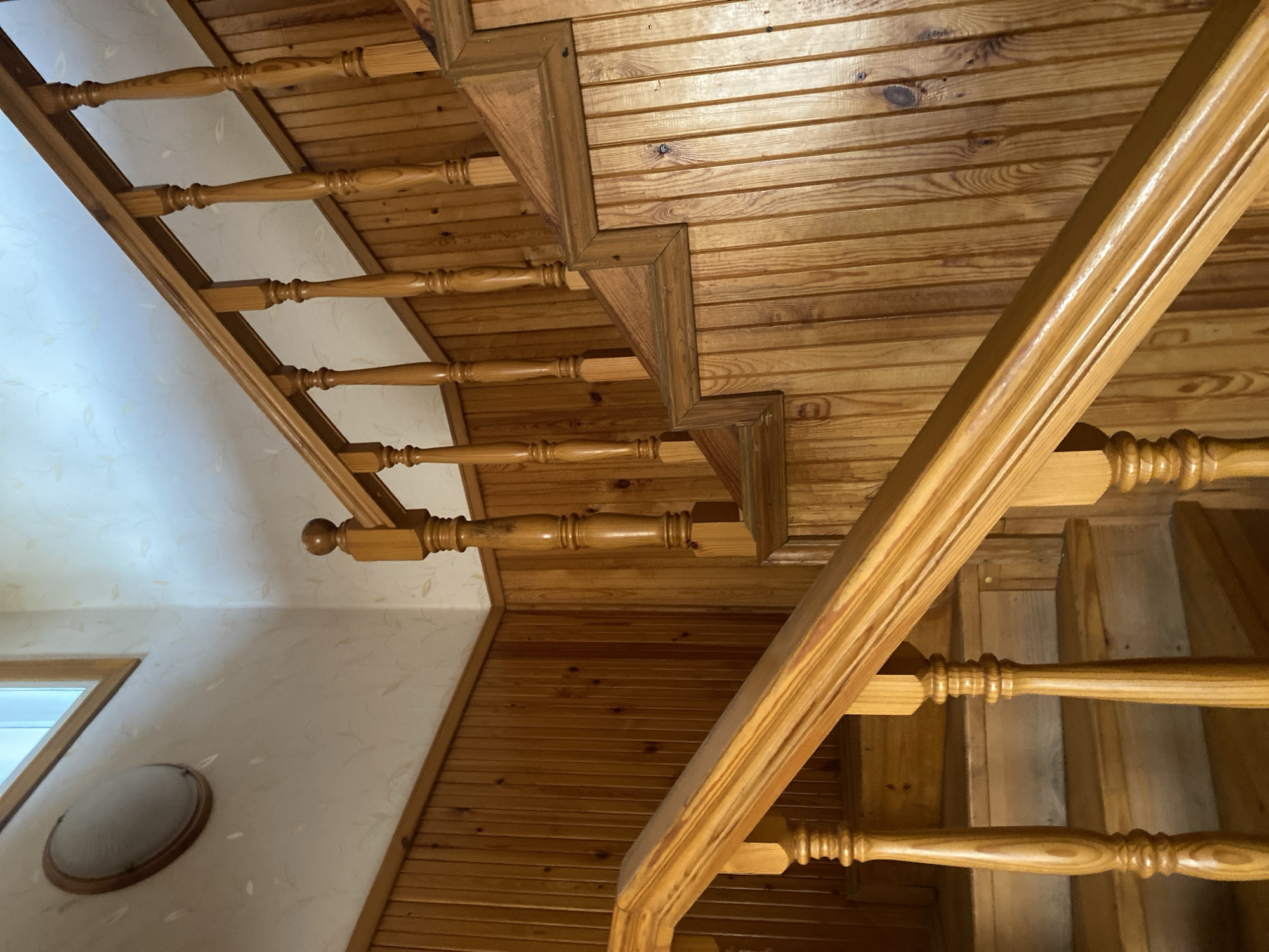 The image shows an interior staircase made of light-colored wood, likely pine or a similar softwood. The walls are paneled with vertical wooden boards up to a certain height, and the upper portion of the walls is covered with light-colored wallpaper featuring a subtle floral pattern. A wooden handrail with turned balusters lines the staircase. There is a round light fixture with a wooden frame on the upper left wall, near a window with a wooden frame. The overall impression is of a rustic, traditional interior. The image is a photograph taken from a low angle, looking up the staircase. There are no people or animals visible in the image. The lighting appears to be natural, coming from the window. The scene is well-lit and the details of the wood grain are visible.