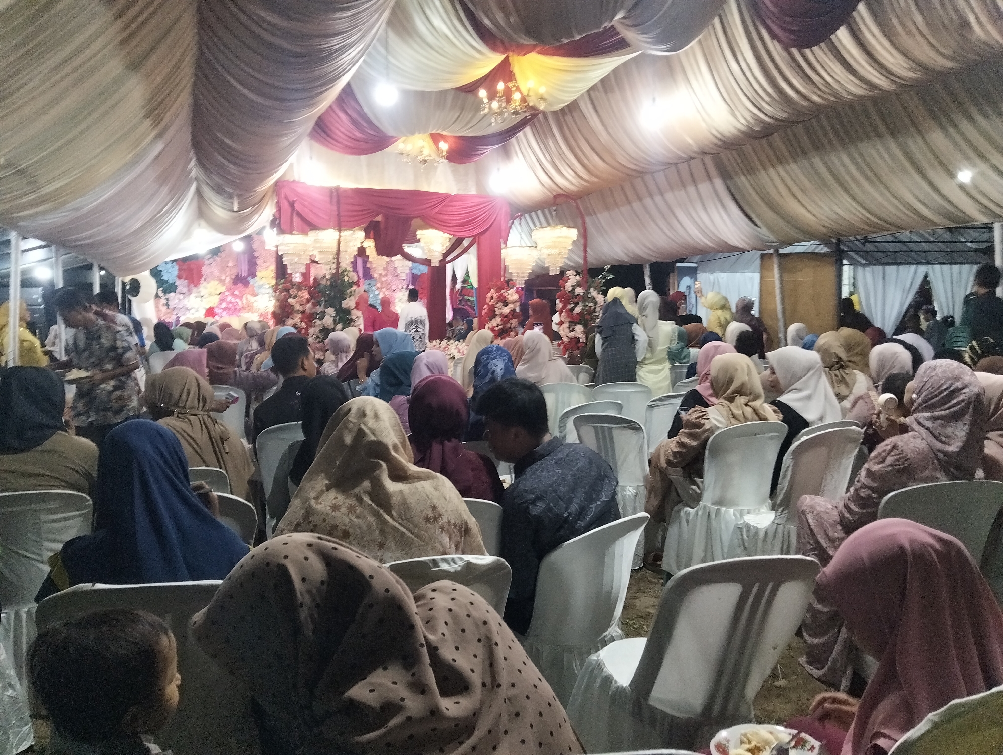 The image depicts an indoor gathering, likely a wedding reception or similar event, under a draped tent ceiling. The tent is decorated with white and maroon fabric, creating a festive atmosphere. Numerous people are seated on white chairs, facing towards a decorated backdrop that includes floral arrangements and draped fabric. The audience is diverse, with men, women, and children present. Many individuals are wearing traditional attire, including hijabs. In the foreground, a young child is visible, looking towards the right side of the frame. The setting appears to be informal, possibly outdoors or in a large open space covered by the tent. The lighting is subdued, with some chandeliers and overhead lights providing illumination. The overall impression is of a community celebration. This event is taking place in Bambalemo, Indonesia.