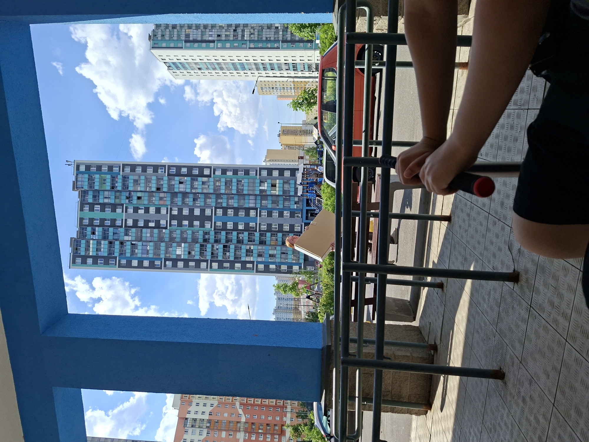 This image captures an urban scene from a sheltered vantage point on a bright, sunny day, likely in Saint Petersburg, Russia. The perspective is from behind a dark metal railing and within a structure framed by prominent blue beams.
In the immediate foreground, on the right, the arms and hands of a person are visible, gripping the red-tipped handles of the railing. The person is wearing dark shorts, and the ground surface is a light-colored, textured pavement with a diamond pattern, upon which strong shadows from the railings are cast.
Beyond the railing and blue frame, a wide expanse of clear blue sky with fluffy white cumulus clouds dominates the background. Several tall residential buildings are visible. A striking high-rise building with a pattern of dark grey, blue, and black panels is prominent on the left. Another tall building, predominantly white with grey sections, stands in the upper right. Further in the distance, numerous other multi-story residential buildings in shades of beige, orange, and light grey are scattered across the landscape, interspersed with green trees.
In the middle ground, a red car is partially visible, positioned behind the railings. A person is also seen holding a large, light-colored rectangular board or panel near the red car. The overall setting is a modern residential area under pleasant, sunny weather conditions. No legible text is visible in the image.