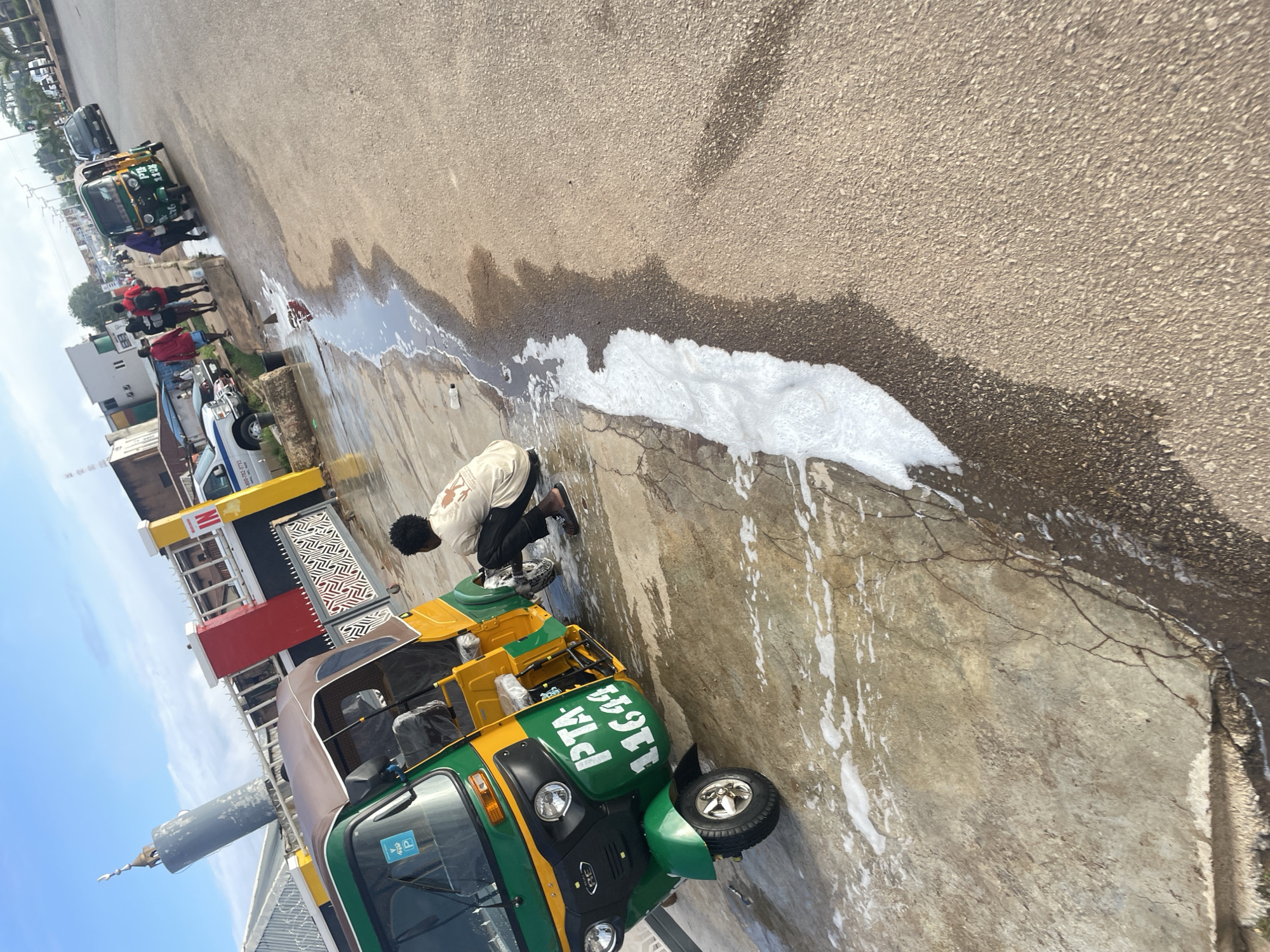 In Barakin Lerawi, Nigeria, a person is seen crouching by a green and yellow auto-rickshaw, engaged in cleaning its wheel. The auto-rickshaw has the license plate "PTA 11644" clearly visible on its front. The ground is wet and foamy, indicating recent washing or a spill. In the background, other vehicles, including a few auto-rickshaws and a white van, are parked or driving along the road. Buildings with various architectural styles are visible along the street, and a tall, cylindrical structure, possibly a minaret, stands out against the sky. The sky is bright and clear, suggesting it is daytime.