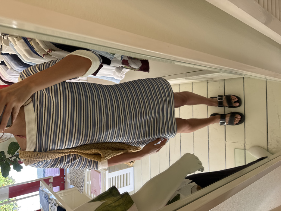 The scene is a retail clothing store in Marina Maroma, Mexico. A person is visible from the upper chest to the feet, reflected in a mirror while taking a smartphone selfie, which obscures their face. The individual wears a boat-neck, short-sleeved dress with horizontal stripes in white, light blue, and thin dark blue. The dress features a white ribbed collar and white ribbed sleeve cuffs. A red and white patterned strap is visible at the left shoulder. A light brown, loosely woven tote bag is slung over the individual's left shoulder. They wear black sandals with two silver-buckled straps on their feet. The person stands on a light-colored wooden plank floor. To the left, a white mannequin arm displays a garment with a green foliage pattern over dark fabric. The background includes retail fixtures: a white slatted display table on the left with folded clothing items, and multiple clothing racks laden with various shirts and blouses in assorted colors and patterns. A green potted plant is partially visible in the upper left background.