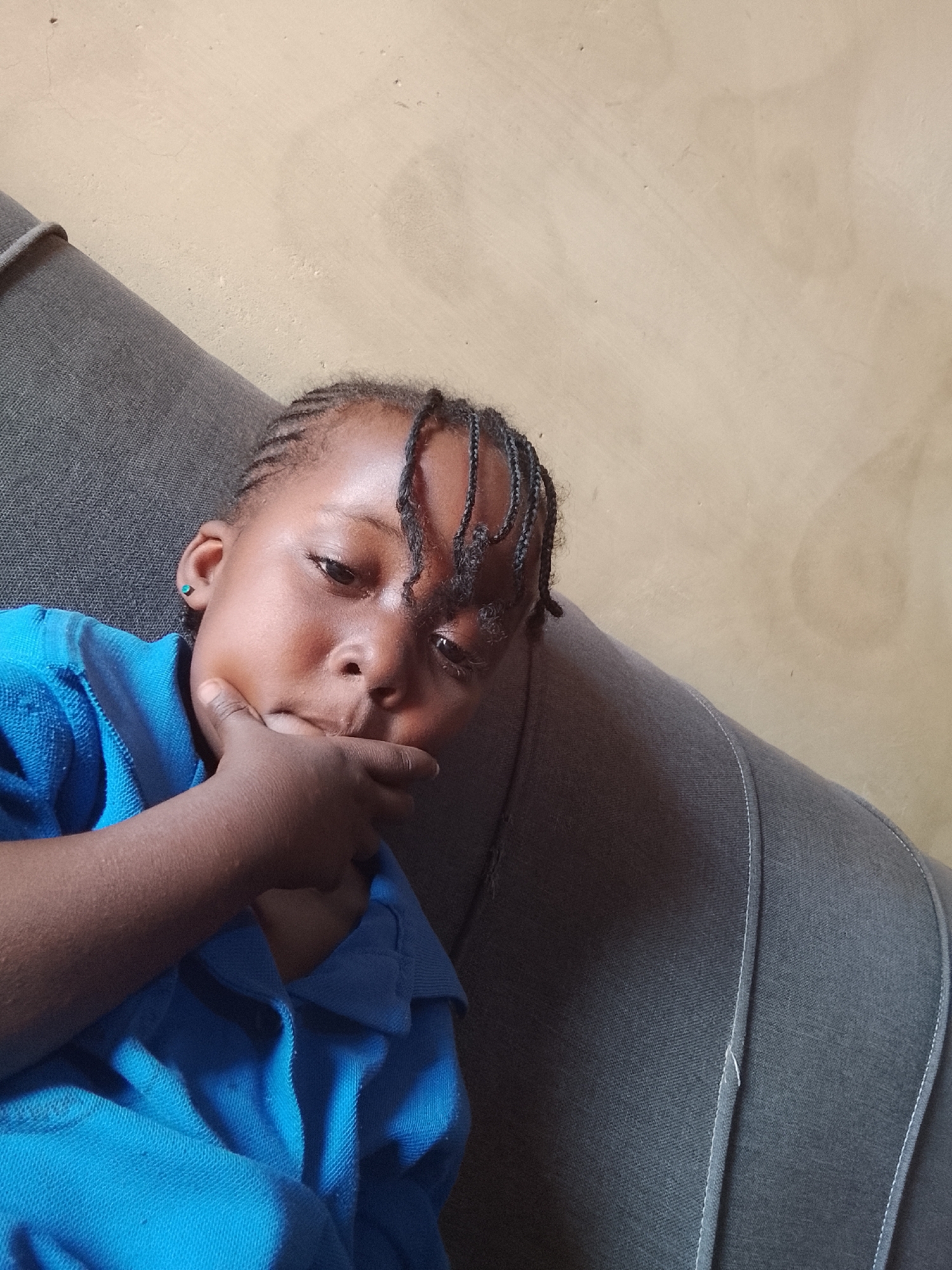A medium close-up, slightly Dutch-angled indoor photograph captures a young child resting on a dark gray sofa. The child, who appears to be of African descent, is positioned in the left half of the frame, looking towards the viewer with a thoughtful or contemplative expression. They are wearing a bright blue short-sleeved top with a collar.
The child's dark hair is styled in neat braids, with some strands falling across their forehead. A small, light blue stud earring is visible in their left earlobe. Their right hand is brought up to their face, with fingers gently curled against their chin and mouth area, reinforcing a pensive mood.
The background features a plain, light beige or tan wall with a slightly textured finish. The child is reclining on a dark gray sofa or armchair, which has distinctive white stitching along its edges and seams. The lighting suggests an indoor daytime setting. The scene depicts a quiet moment in an indoor environment, consistent with a home in Kuru Dungus, Nigeria. There is no visible text in the image.
