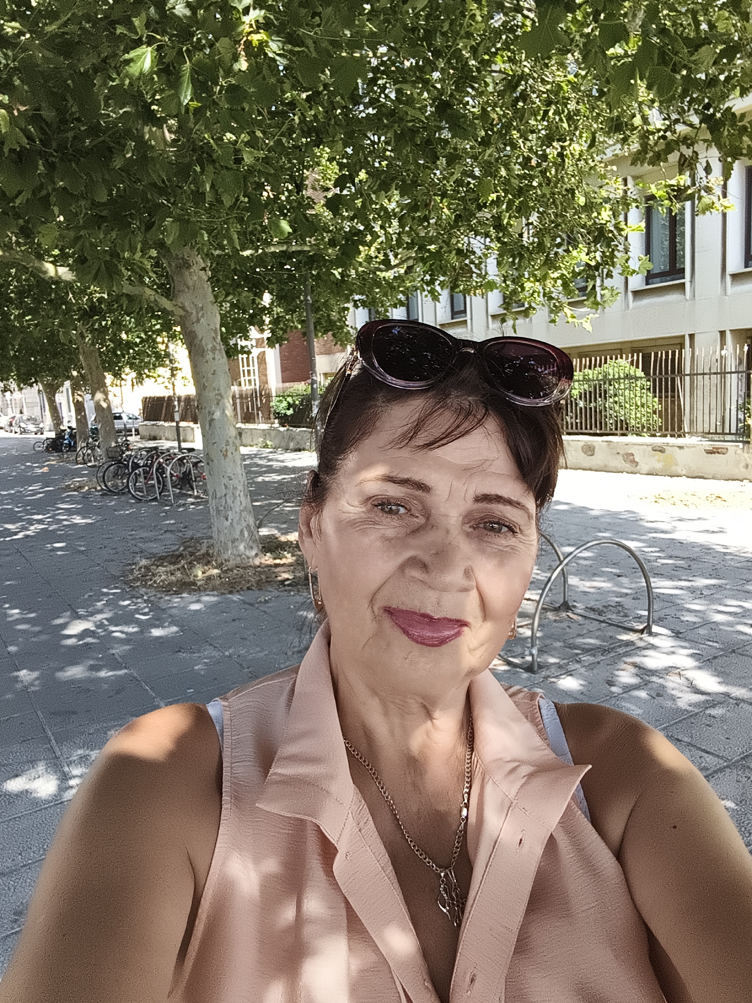 This is a medium shot, eye-level selfie of a woman taken outdoors on a sunny day. The woman has brown hair styled up and is wearing sunglasses on top of her head. She has a light peach-colored sleeveless collared shirt on with a silver necklace. Her lips are painted a bright pink. She appears to be smiling faintly. The background shows a sidewalk lined with trees, casting dappled shadows. Beyond the trees, there is a building with windows and a fence. Several bicycles are parked in racks in the background. The presence of bicycles and the outdoor setting suggest a leisurely activity. The bright sunlight indicates it is daytime. The architecture and parked bicycles are consistent with a European setting, possibly a city or town.