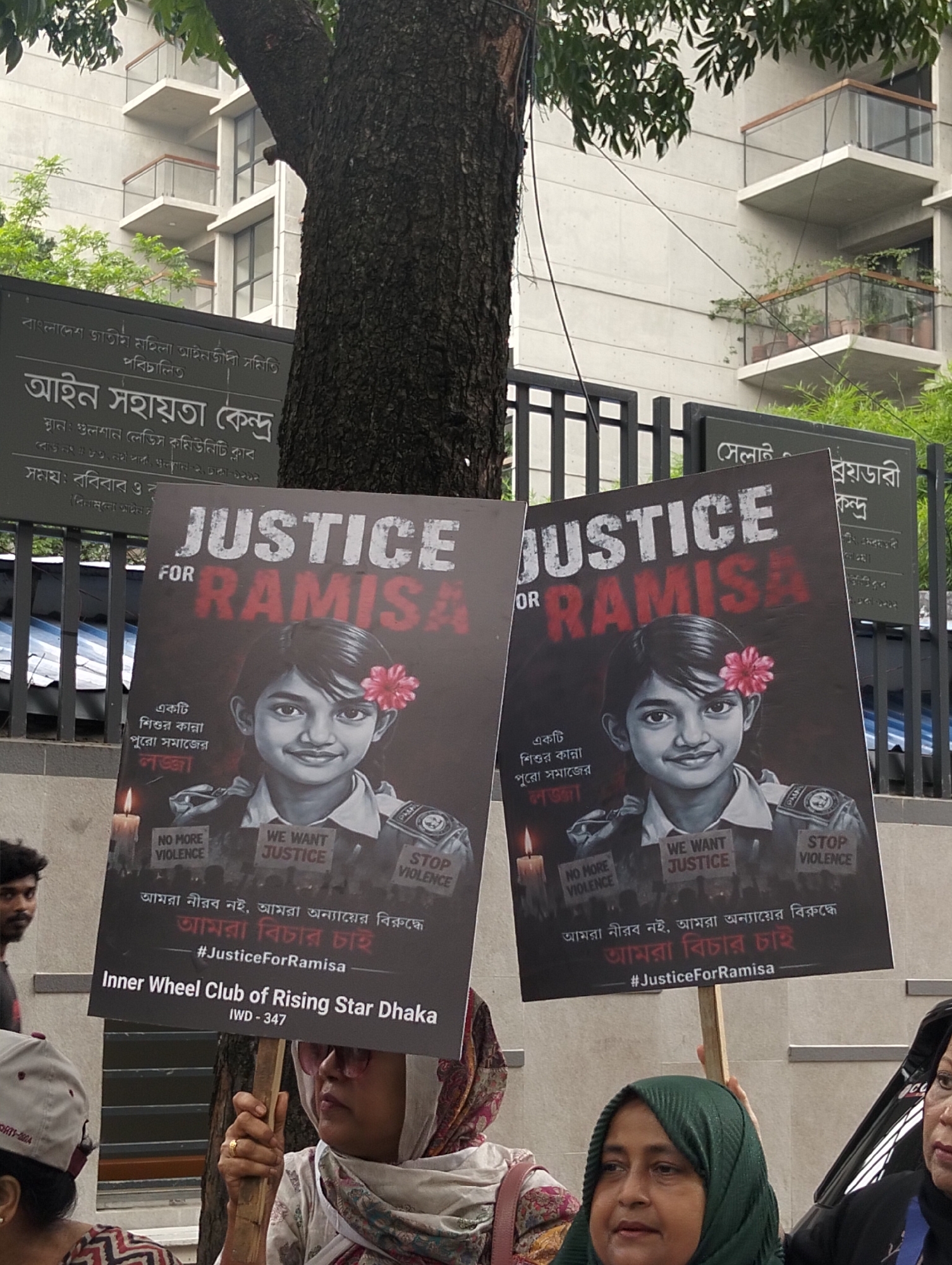 The image shows two signs being held up at a protest in Dhaka, Bangladesh, likely near the "Gulshan Ladies Community Club" (as indicated by the nearby sign "আইন সহায়তা কেন্দ্র" or "Legal Aid Center"). The protest signs read "JUSTICE FOR RAMISA," and feature a portrait of a young girl with a flower in her hair. Below the portrait, the signs state "একটি শিশুর কান্না পুরো সমাজের লজ্জা," which translates to "A child's cry, the shame of the whole society." The signs also include the phrases "NO MORE VIOLENCE," "WE WANT JUSTICE," and "STOP VIOLENCE." The bottom of the signs indicates the protest is being organized by the "Inner Wheel Club of Rising Star Dhaka." Several people are visible, including women holding the signs, and a man in the background.