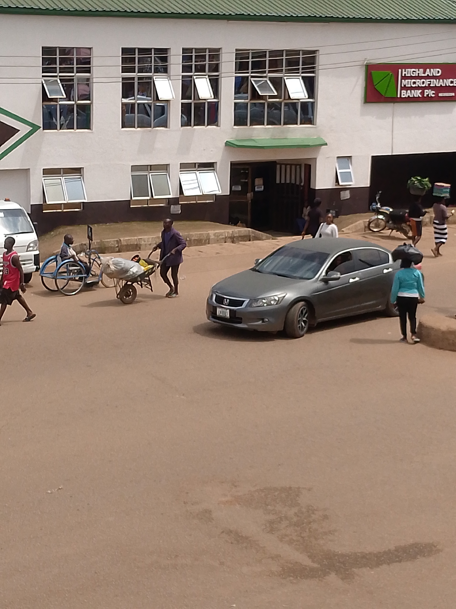 In the city of Bukuru, Nigeria, a street scene unfolds outside a building identified as Highland Microfinance Bank Plc. The building features a white facade with multiple windows, some of which are open. A prominent green and maroon sign indicates the bank's name.
In the foreground, a gray sedan is parked. A woman in a blue top and black pants walks past the car, carrying a load on her head. To the left of the car, a man in a red vest and black shorts walks away from the camera.
Further left, two men are interacting near a wheelchair. One man, in a purple top and dark pants, is pushing a wheelbarrow loaded with what appears to be sacks. The other man is seated in the wheelchair, which has large spoked wheels. A white van is parked behind them.
The ground is paved with asphalt. In the background, a motorcycle is parked near the bank's entrance. Other individuals are visible near the bank, some walking and others standing. The overall environment suggests a busy commercial area.