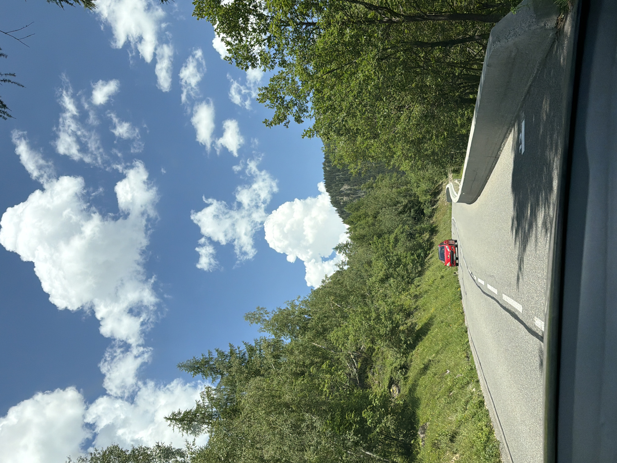 The image captures a view from a vehicle on a winding mountain road in Vallorcine, France. The road is paved and marked with white lines. A red car is driving ahead on the road. The road is bordered by a concrete barrier on the right and a grassy slope with trees on the left. The surrounding environment is lush with green trees and vegetation, suggesting a natural, mountainous area. The sky is blue with scattered white clouds. The scene is likely a scenic route within the French Alps.