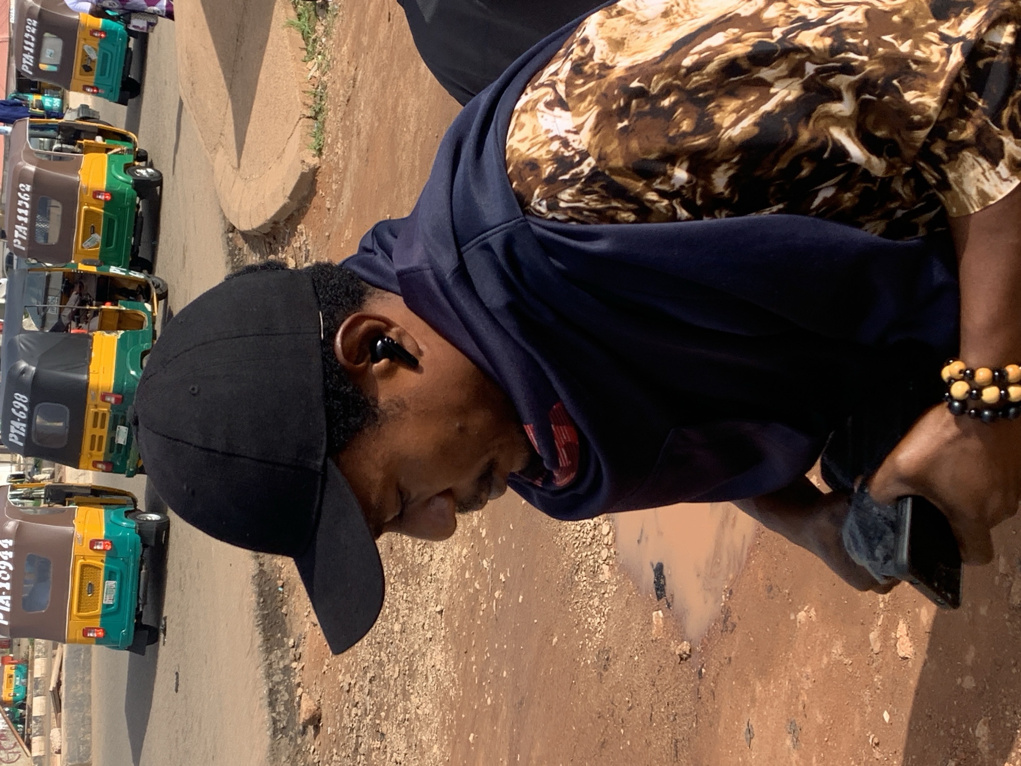 A man wearing a black baseball cap, a navy blue hoodie, and a brown and white patterned shirt is standing outdoors in what appears to be a street. He is holding a smartphone in his right hand. He has an earpiece in his left ear. In the background, there are several auto-rickshaws, some of which have license plates that begin with "PTA." The ground is a mix of dirt and small stones, with a small puddle of murky water to the left of the man. The overall scene suggests a busy street environment, possibly in Nigeria given the license plate prefixes.