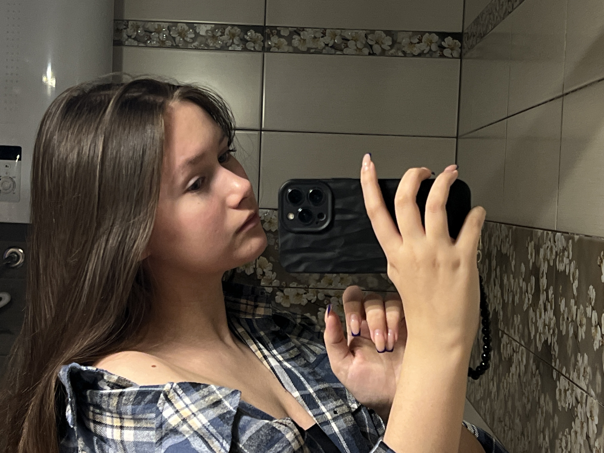 A young woman is depicted taking a mirror selfie indoors, looking towards her left with a neutral to slightly pensive expression. She has long, straight, dark brown hair and is wearing a blue and white plaid shirt, pulled down off her shoulders. She holds a black smartphone, featuring a triple camera array, in her right hand to capture the reflection. Her fingernails are adorned with a French manicure style, tipped in a dark blue or purple color. A black beaded strap is attached to the phone. A small mole is visible on her right collarbone area. The background consists of light-colored, possibly beige or grey, rectangular wall tiles, accented by a decorative floral border with white flowers on a darker background running horizontally. On the far left of the image, a white appliance with a circular dial and buttons is partially visible, suggesting a bathroom or utility area. The scene is illuminated by artificial lighting. Based on the provided context, the inferred location is Chaikovsky, Russia. There is no visible text in the image.