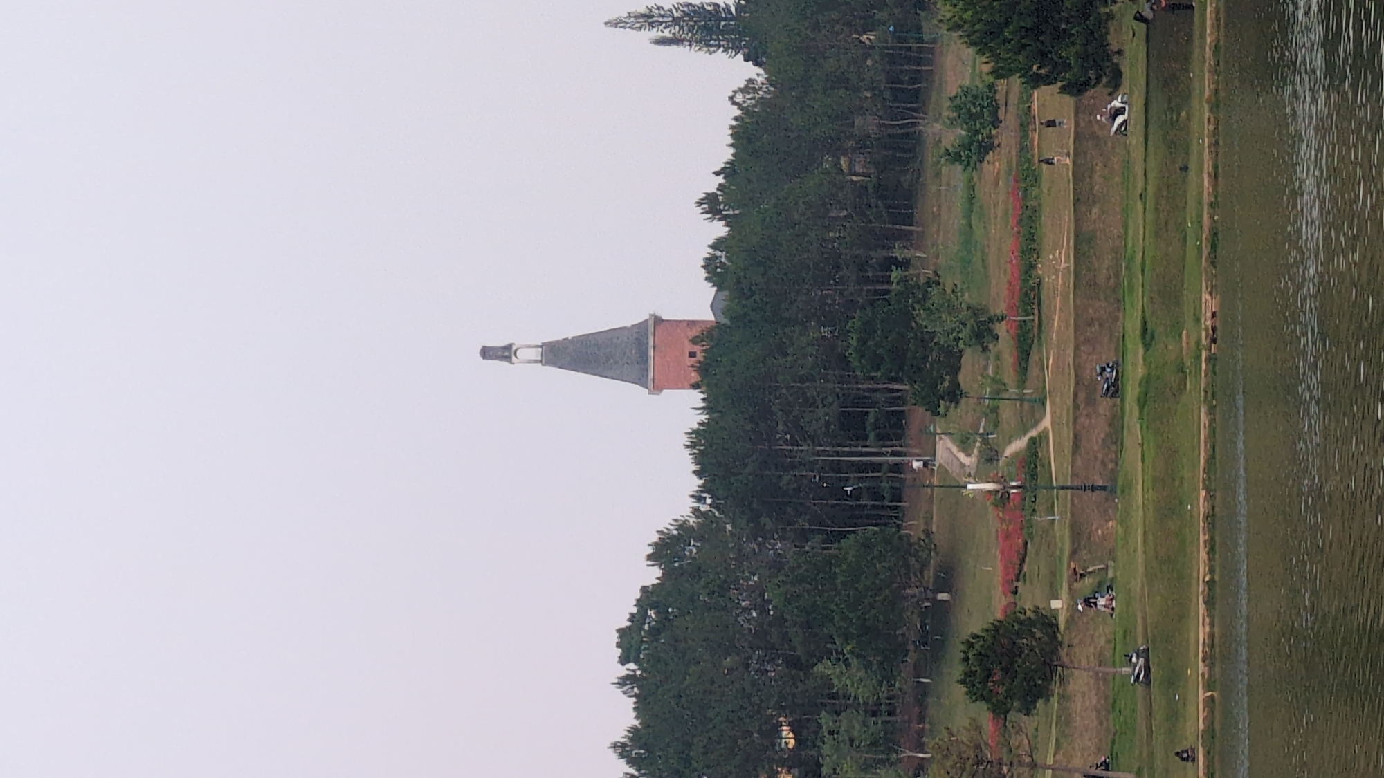 The image shows a view of Da Lat, Vietnam. A tall structure, likely a church or bell tower with a conical roof, rises above the trees in the center. Adjacent to this is a long and narrow lake or pond running alongside a grassy, park-like area with pedestrian paths. There are several figures walking or sitting near the lake, and some vehicles are parked. The sky is overcast and gray, suggesting a hazy or foggy atmosphere.