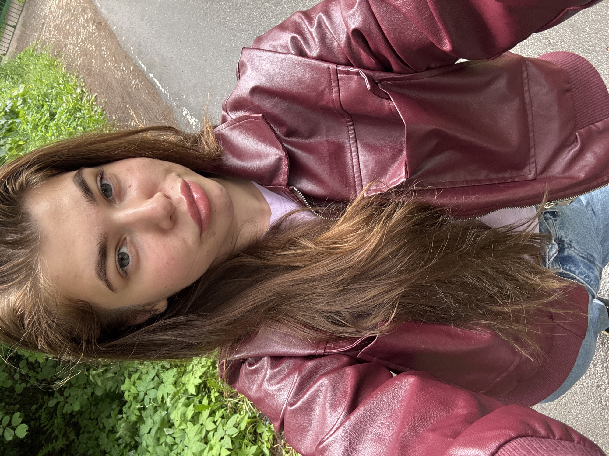 A young woman with long, brown hair is taking a selfie outdoors. She is wearing a burgundy faux leather jacket over a pink t-shirt and blue jeans. Her lips are full and appear to be glossed. The background consists of greenery and a paved path. The scene is likely located on a street or park path within the city of Мещёры, Russia.