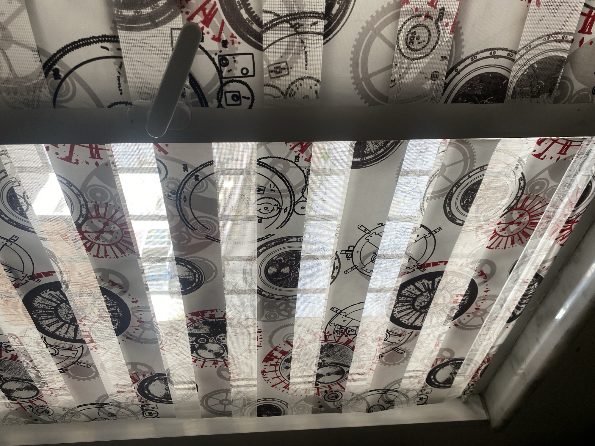 This is a close-up, eye-level shot of a zebra blind with a mechanical-themed pattern. The blind is partially open, revealing a window with sunlight streaming through. The pattern on the blind features various gears, clock faces, and some red accents, giving it a vintage or steampunk aesthetic. To the right of the blind, a white window handle is visible. The scene appears to be indoors, likely in a room with a window. The specific location in Küçükçekmece, Turkey is not discernible from the visual information alone.