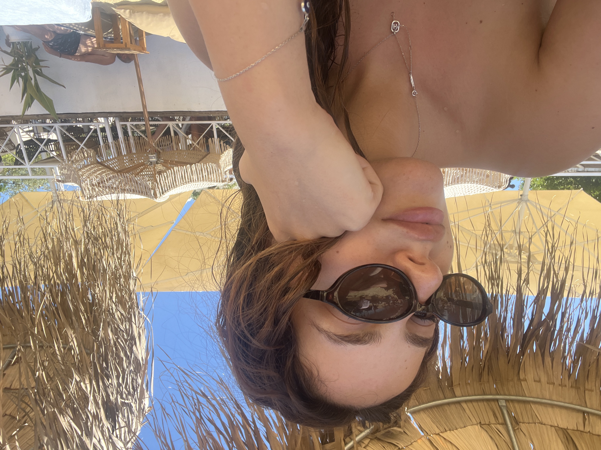 A young woman with dark hair, wearing sunglasses and a bikini, is posing for a selfie. She is sitting under a thatched umbrella on a beach. Her left hand is supporting her chin. In the background, there are more umbrellas, beach chairs, and some people. The scene appears to be in Achladias, Greece.