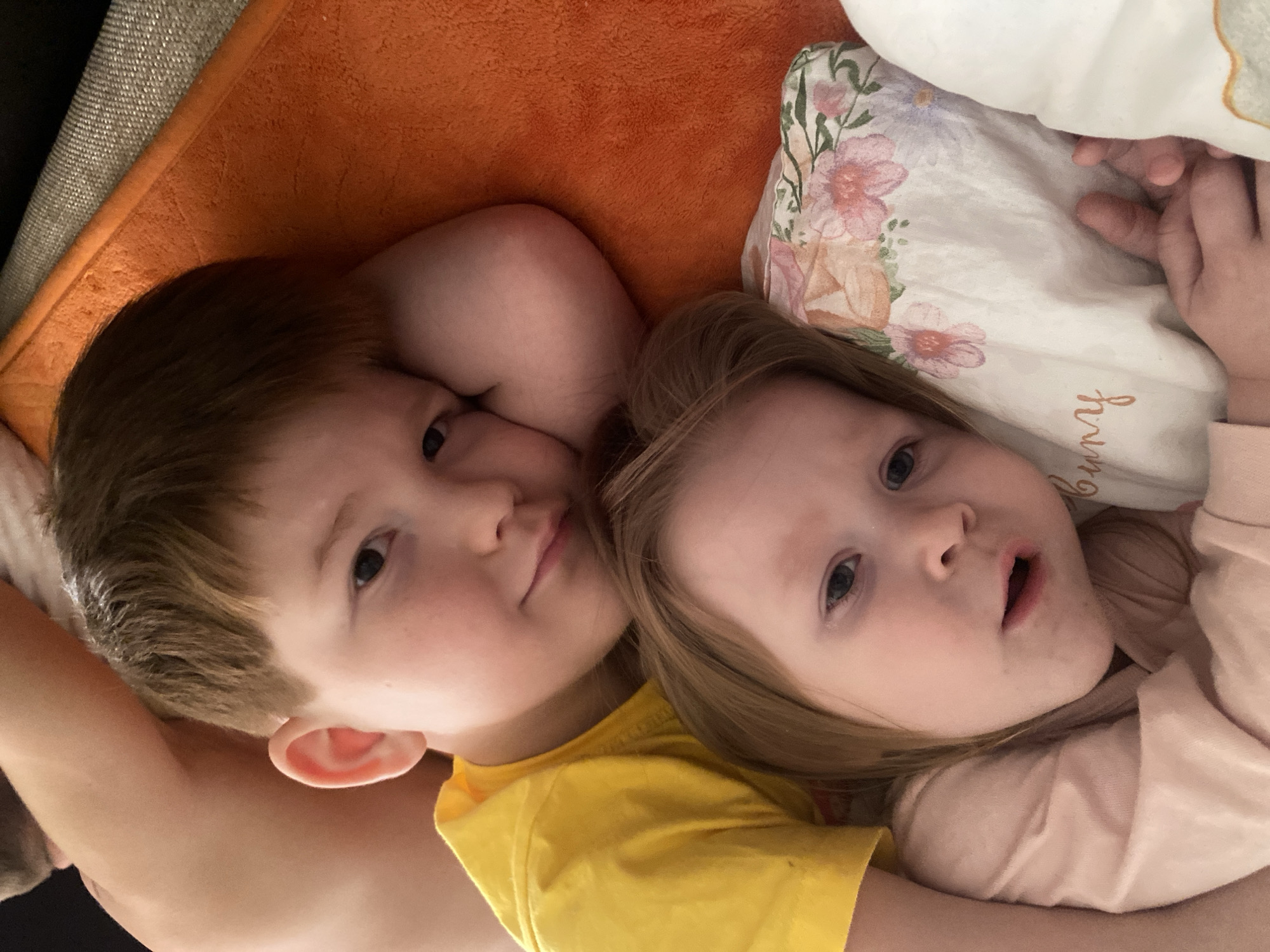 Two young children are lying down next to each other. The child on the left has brown hair and is wearing a yellow shirt. The child on the right has blonde hair and is wearing a light pink shirt. They are lying on an orange surface next to a floral pillow. The location cannot be determined from the image.