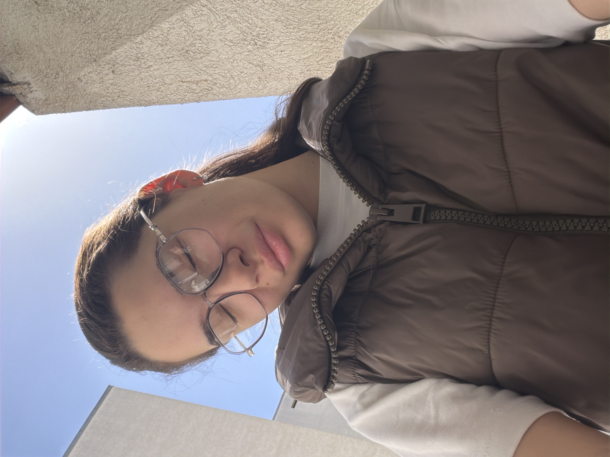 The image displays a low-angle portrait of a young person, rotated 90 degrees clockwise, with their head positioned towards the bottom-right of the frame. The subject is situated outdoors under a bright sky, likely in Sofia, Bulgaria, looking subtly towards the camera with a relaxed expression, featuring slightly parted pink lips and fair skin. They have dark hair and wear prominent round-framed glasses, which have a thin chain attached to the arms and reflect the bright surroundings.
For attire, the individual sports a white long-sleeved top underneath a dark brown puffer vest, characterized by its textured, slightly shiny fabric and a visible zipper running down the front.
The background is dominated by a clear, bright blue sky, indicating a sunny daytime setting. Above the subject's head, a textured, light-colored concrete or stucco overhang or ceiling is visible, suggesting the presence of a building or balcony structure. Another pale building facade is partially seen on the right side of the frame. Strong natural sunlight illuminates the scene, casting reflections on the glasses and creating a subtle glow around the person's hair. No discernible text is visible within the image.