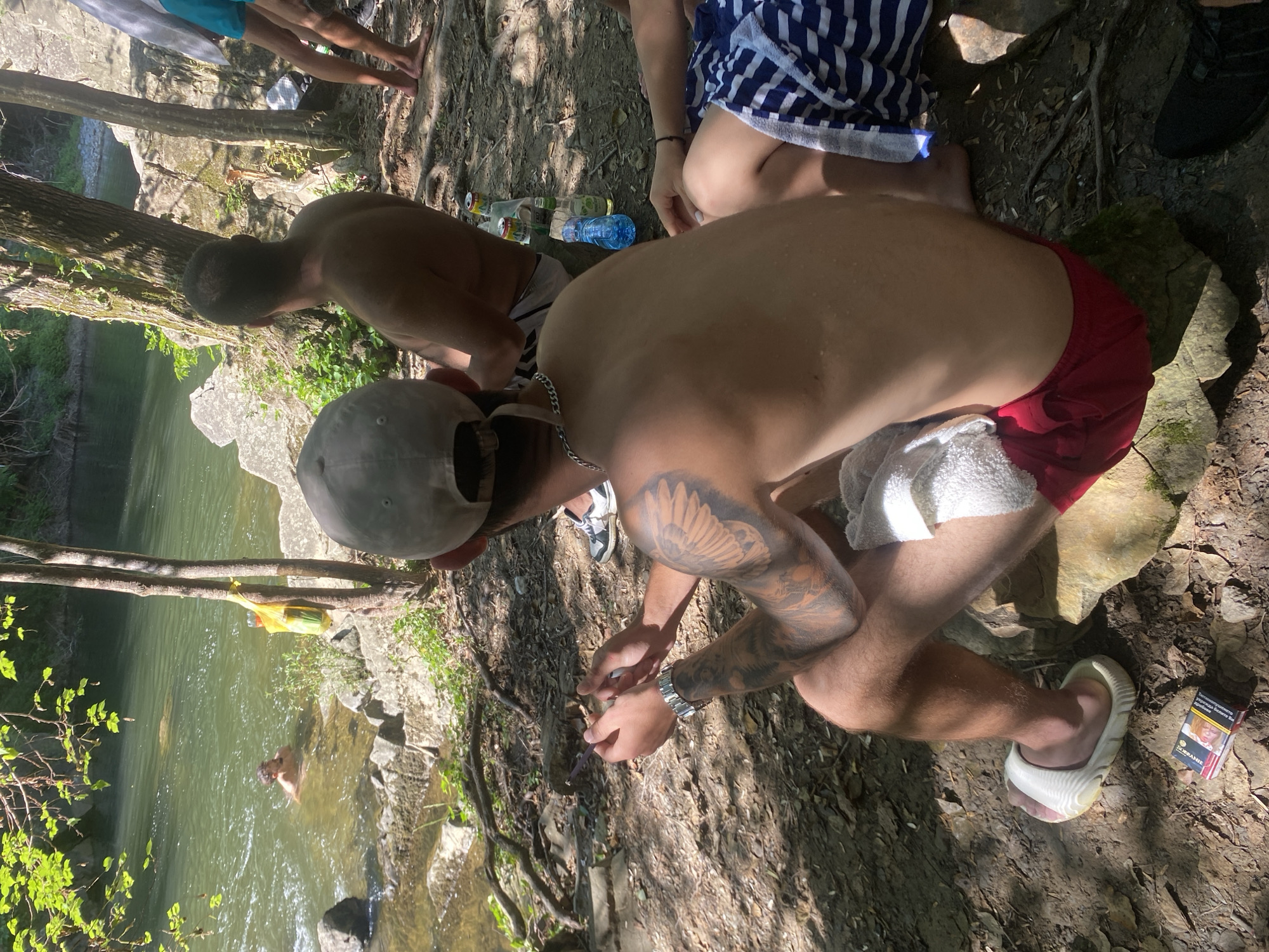 Two young men are gathered by a river in what appears to be a natural, wooded setting, possibly during a warm day. One man, wearing red swim trunks, is partially visible from the side, with a large tattoo of a wing on his arm. He is crouching down, looking at something in his hands. He is also wearing a wristwatch and a white flip-flop on his right foot. A pack of cigarettes is on the ground to his right. The other man, seen from behind, is wearing a grey baseball cap and shorts, and is also by the river. He has his back to the camera, and is leaning forward. In the background, a third person is visible in the water, seemingly swimming or wading. The riverbank is rocky and covered in leaves and tree roots. The sun is shining, casting dappled shadows through the trees, suggesting it is daytime. The overall impression is of a casual outing to a river on a pleasant day. There is some visible text on the cigarette pack, but it is not legible.
