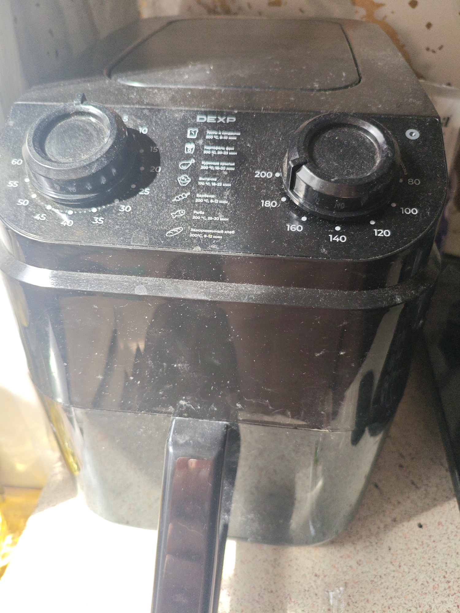 The image shows the top and front of a black DEXP air fryer, heavily coated in dust. The air fryer has two control knobs: one on the left with markings from 5 to 60, and another on the right with temperature markings from 80 to 200 degrees Celsius. A printed guide with icons and text in Russian is visible on the right side of the control panel, detailing cooking times and temperatures for various food items, such as pasta, french fries, chicken wings, and fish. The air fryer is placed on a countertop. The background is indistinct but appears to be an indoor environment. There is no visible indication of the city of Berdsk municipality, Russia, in the image itself.