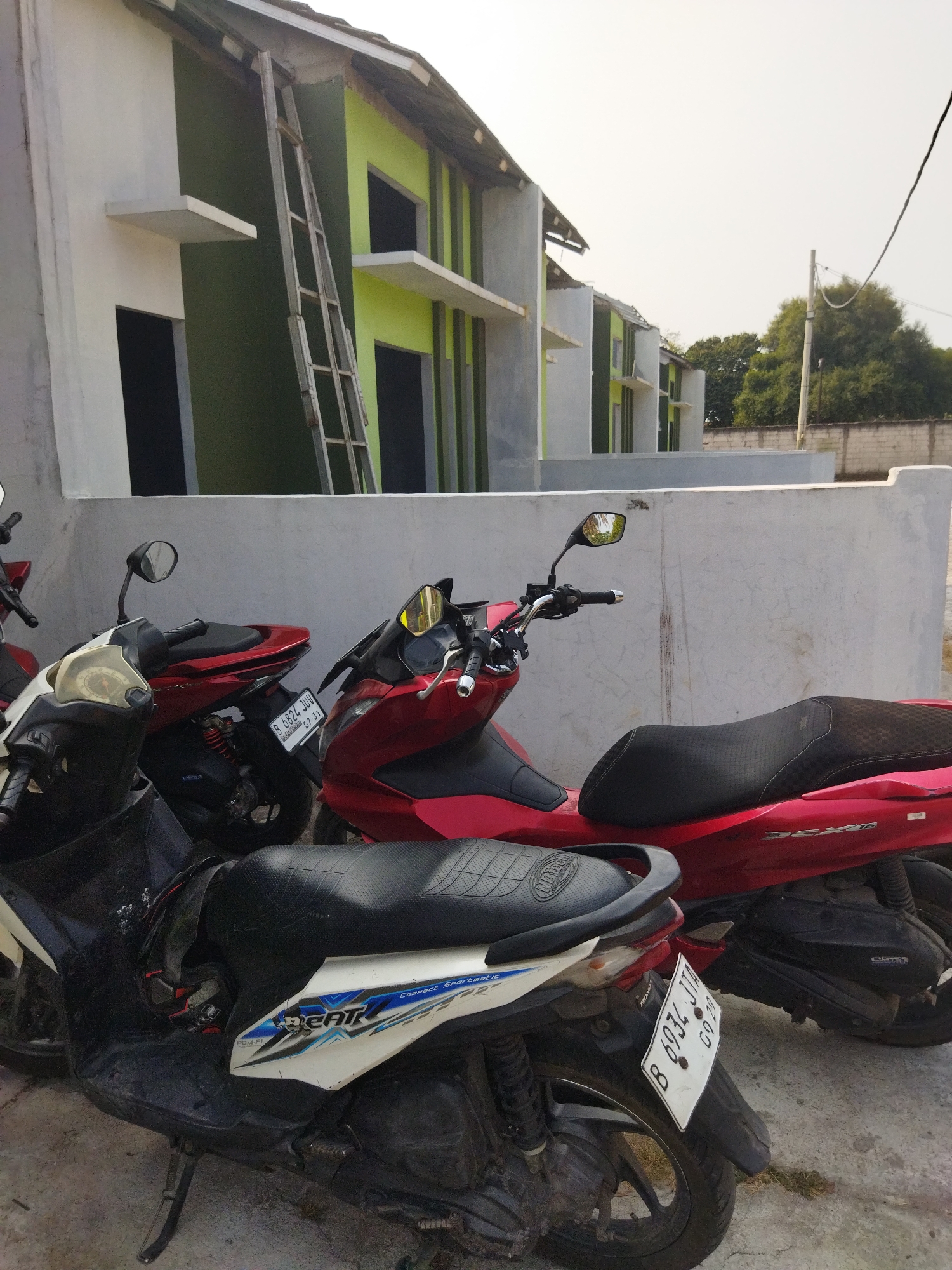 In the city of Suka Sari, Indonesia, a row of houses under construction is visible, painted with contrasting white and lime green exteriors. A tall, aluminum ladder leans against one of the buildings, suggesting ongoing work. In the foreground, three scooters are parked on a concrete surface. The closest scooter is white and black, with the word "BEAT" prominently displayed on its side. To its right, a bright red scooter is parked, and further behind it, another red scooter is partially visible. The scene appears to be an outdoor residential area, possibly a new housing development, with a clear sky overhead.
