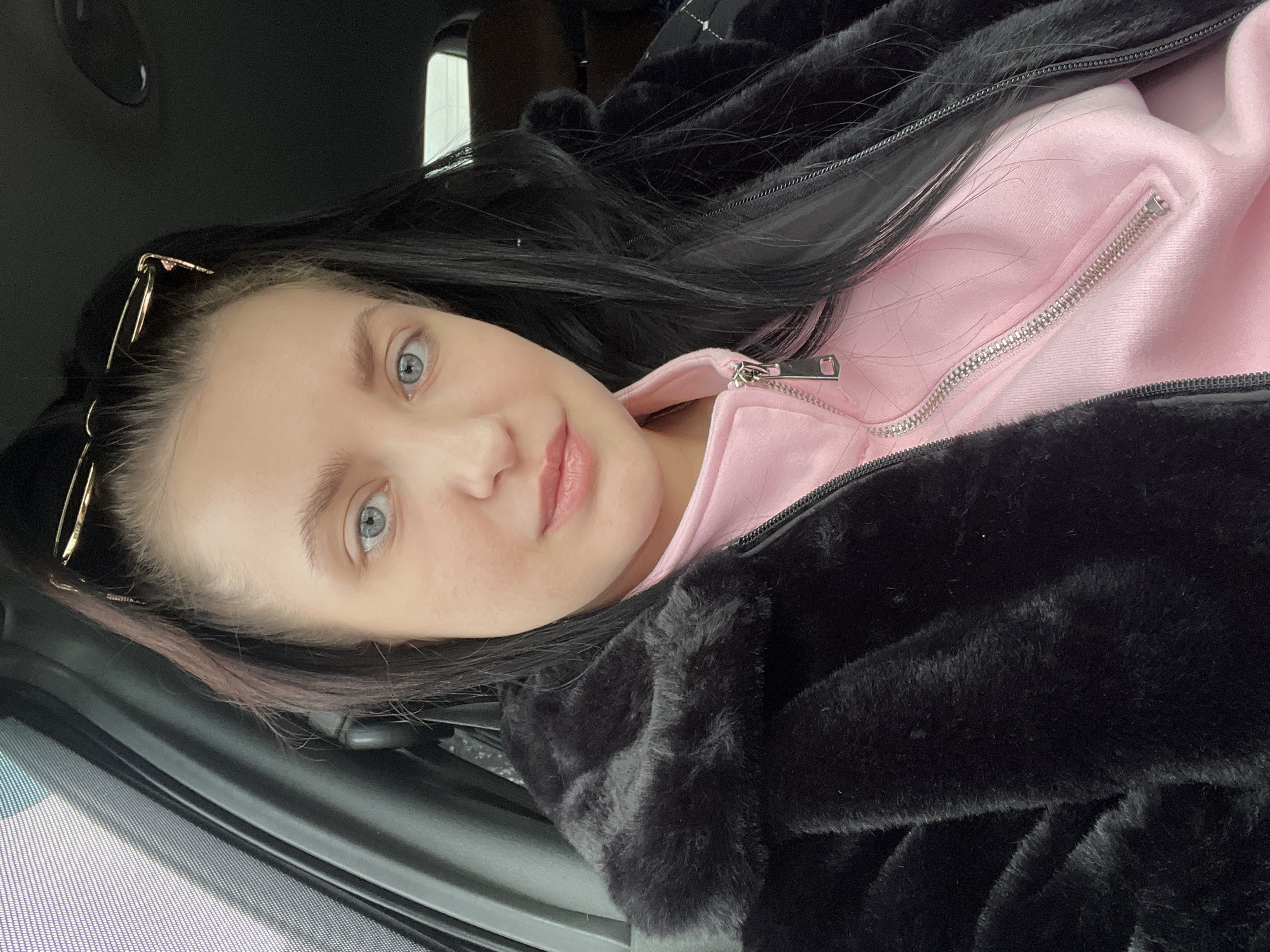 The image presents a close-up, eye-level view of a young adult female seated inside a vehicle, captured as a self-portrait. She has fair skin, prominent blue eyes, and long, dark hair styled partially up, with sunglasses perched on top of her head. Her lips are lightly colored, and she exhibits a neutral to slightly smiling expression, gazing directly at the camera. She is wearing a black faux fur coat over a light pink zippered top, which features a silver zipper with a distinct rectangular pull tab. The background reveals elements of a car's interior, including a dark headliner above and a dark-colored pillar on the left, partially obscuring a window with a fine mesh pattern. Diffused daylight illuminates the scene from the left. This event is stated to occur in the city of Chaikovsky, Russia.