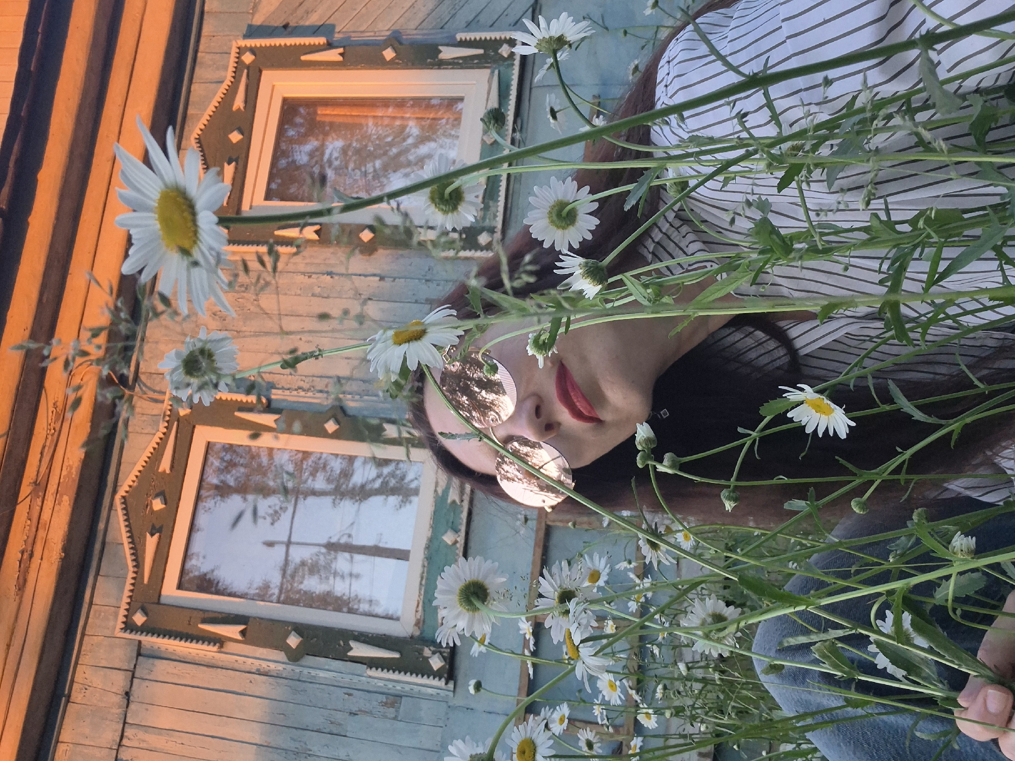 The image depicts a low-angle outdoor shot focusing on a woman amidst a field of daisies in front of a weathered blue wooden house. The woman, with dark hair and wearing round, rose-gold tinted sunglasses and a striped shirt, is positioned slightly off-center to the right. Her face is tilted upwards, and she is looking towards the right side of the frame. Her lips are painted red. The house, painted in faded blue planks, features two decorative windows with intricate white and dark green trim. The upper portion of the house is illuminated by the warm, orange glow of what appears to be sunset or sunrise. The daisies, with white petals and yellow centers, are scattered throughout the foreground, with some stems and leaves obscuring parts of the woman and the house. The overall setting suggests a rural or suburban environment, possibly a dacha or a village house, common in the outskirts of cities like Chaikovsky, Russia, known for its natural landscapes and wooden architecture. The lighting creates a nostalgic and peaceful atmosphere.