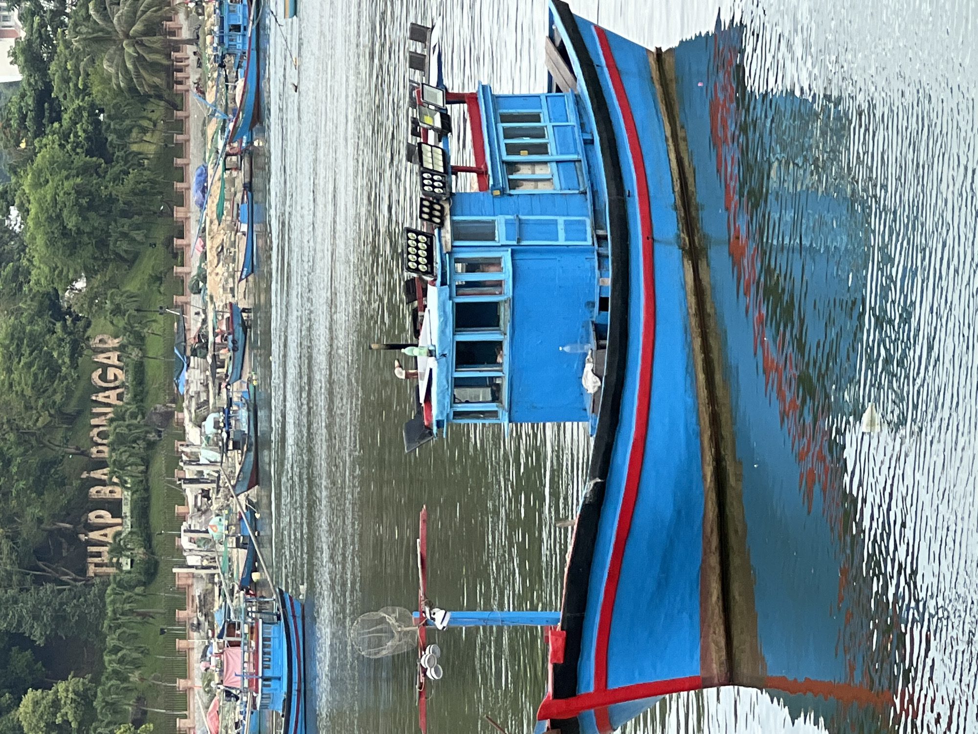 The image depicts a bright blue fishing boat with red trim, moored in a body of water. The boat has a cabin with multiple windows and is equipped with several large spotlights on its roof. In the background, other boats are visible, as well as a shoreline with trees and a sign that reads "THAP BA HON A". This sign is a landmark in Nha Trang, Vietnam, indicating the location is likely near the Thap Ba Hot Springs or the Po Nagar Cham Towers. The water is calm with ripples, reflecting the boat and the surrounding scenery. The overall scene suggests a harbor or a bay area in Vietnam.