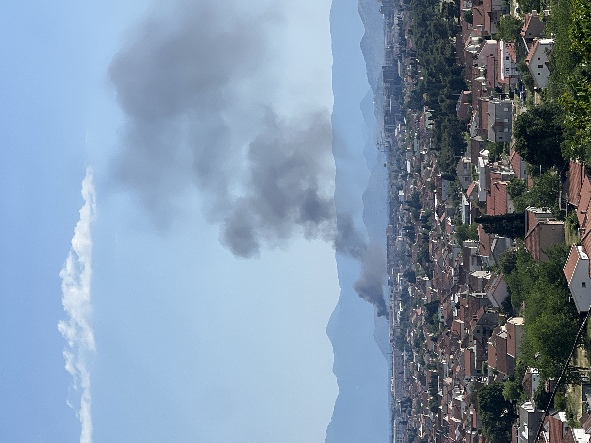 The image shows a view of a city, presumably Podgorica, Montenegro, with smoke rising into the air. A dense residential area of tightly packed buildings with red-tiled roofs dominates the lower portion of the image, leading into taller buildings and cityscape in the mid-ground. A backdrop of hazy blue mountains lies behind the city. Thick, dark gray smoke billows upwards towards the left side of the frame, suggesting a fire or other combustion event occurring within the city. The sky is a mix of clear blue and lighter, diffuse clouds.