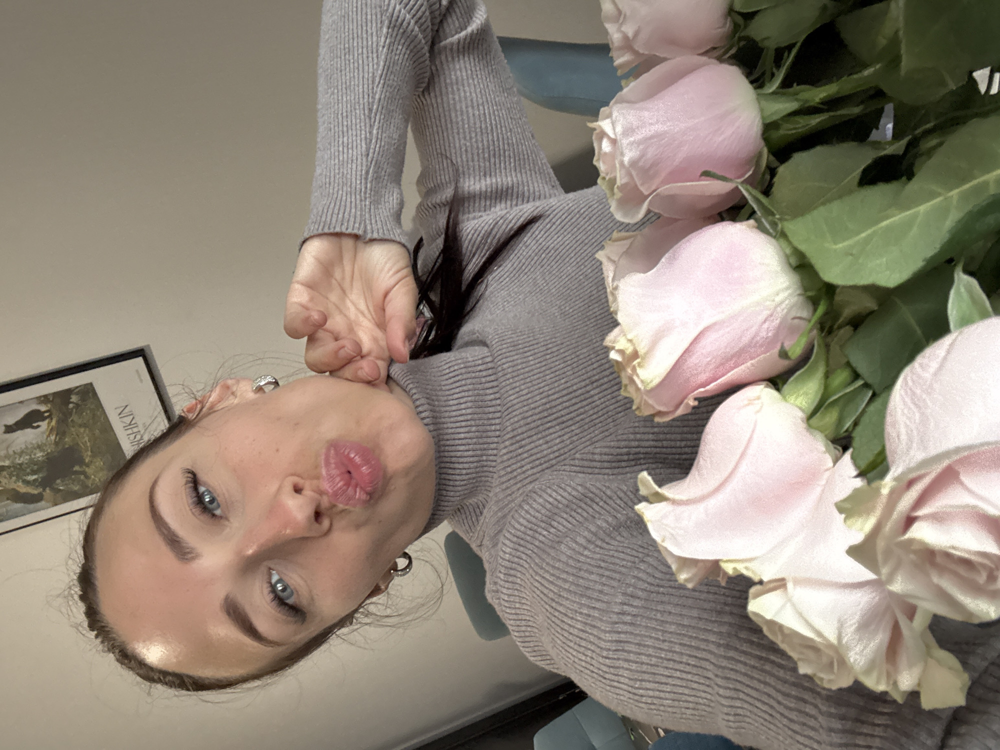A woman with blue eyes and dark hair is posing with a bouquet of light pink roses. She is wearing a gray turtleneck sweater. Her left hand is near her face, and she is making a kissing face. Behind her is a framed picture on the wall. The location is suggested to be Saint Petersburg, Russia, but there is no evidence of this in the image.