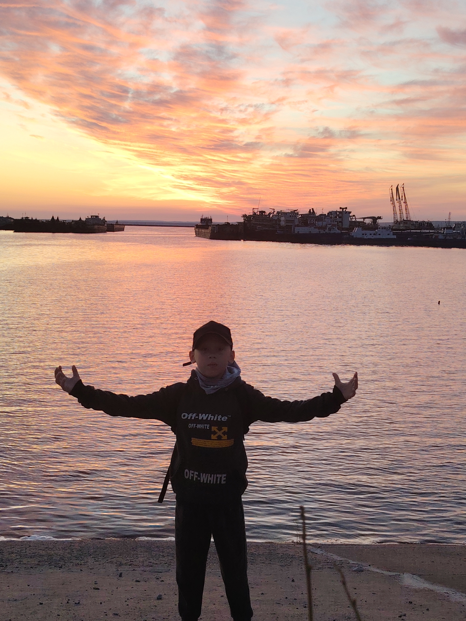 A young boy stands centered in the foreground of a waterfront scene, facing the camera with his arms outstretched and palms slightly upturned. He wears a dark baseball cap and a black hoodie, prominently featuring the "OFF-WHITE™" brand name in white text across the chest and the lower body, along with the brand's yellow diagonal arrows logo on the chest.
The scene is set during either sunrise or sunset, indicated by the dramatic and vibrant sky. Streaks of orange, pink, yellow, and light blue clouds fill the sky, casting a warm, soft glow across the entire image. This golden light is reflected beautifully on the calm surface of the water, creating a mirror effect of the sky's colors.
In the background, across the wide body of water, several large ships or barges are visible, silhouetted against the bright horizon. Further in the distance, industrial cranes and structures suggest a working port or a dockside area. The boy is standing on a concrete or dirt bank, with the water gently lapping at the edge. The overall atmosphere is serene and picturesque, highlighting the natural beauty of the sky and water.
The location context suggests this scene is along the Kama River in Chaikovsky, Russia.