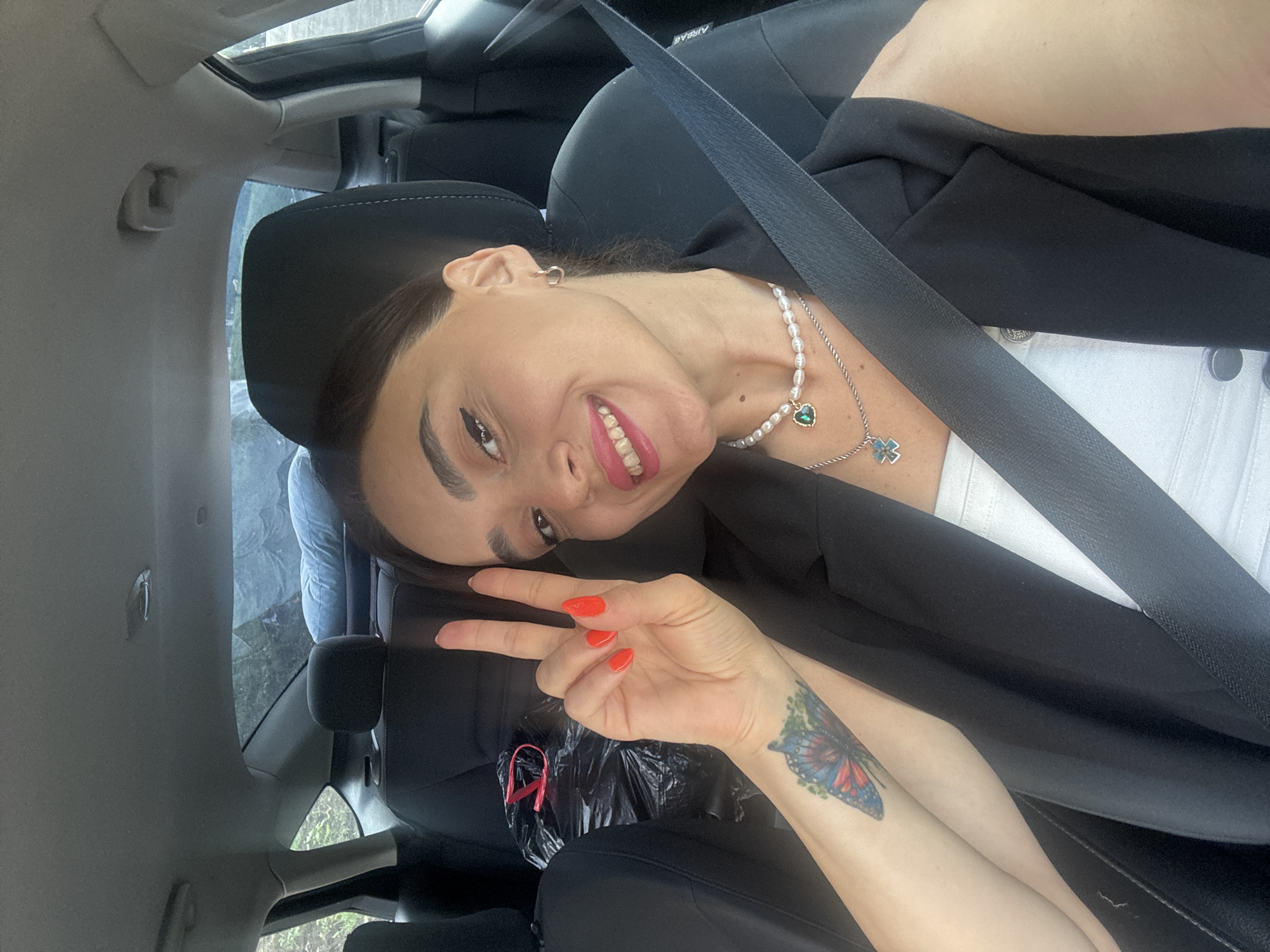 A young woman with dark hair and dark eyes is sitting in the passenger seat of a car. She is wearing a white top, a black blazer, and a pearl necklace with a green heart pendant. Her right hand is raised, and she is holding up two fingers in a peace sign. She has bright red nail polish. A butterfly tattoo is visible on her left forearm. The car's interior is visible, including the black seats, seatbelt, and the headrest of the driver's seat. A black plastic bag with a red strap is on the back seat. The background outside the car windows shows trees and foliage, suggesting a natural or semi-rural setting. The overall lighting suggests daylight. The location is identified as Tbilisi, Georgia.