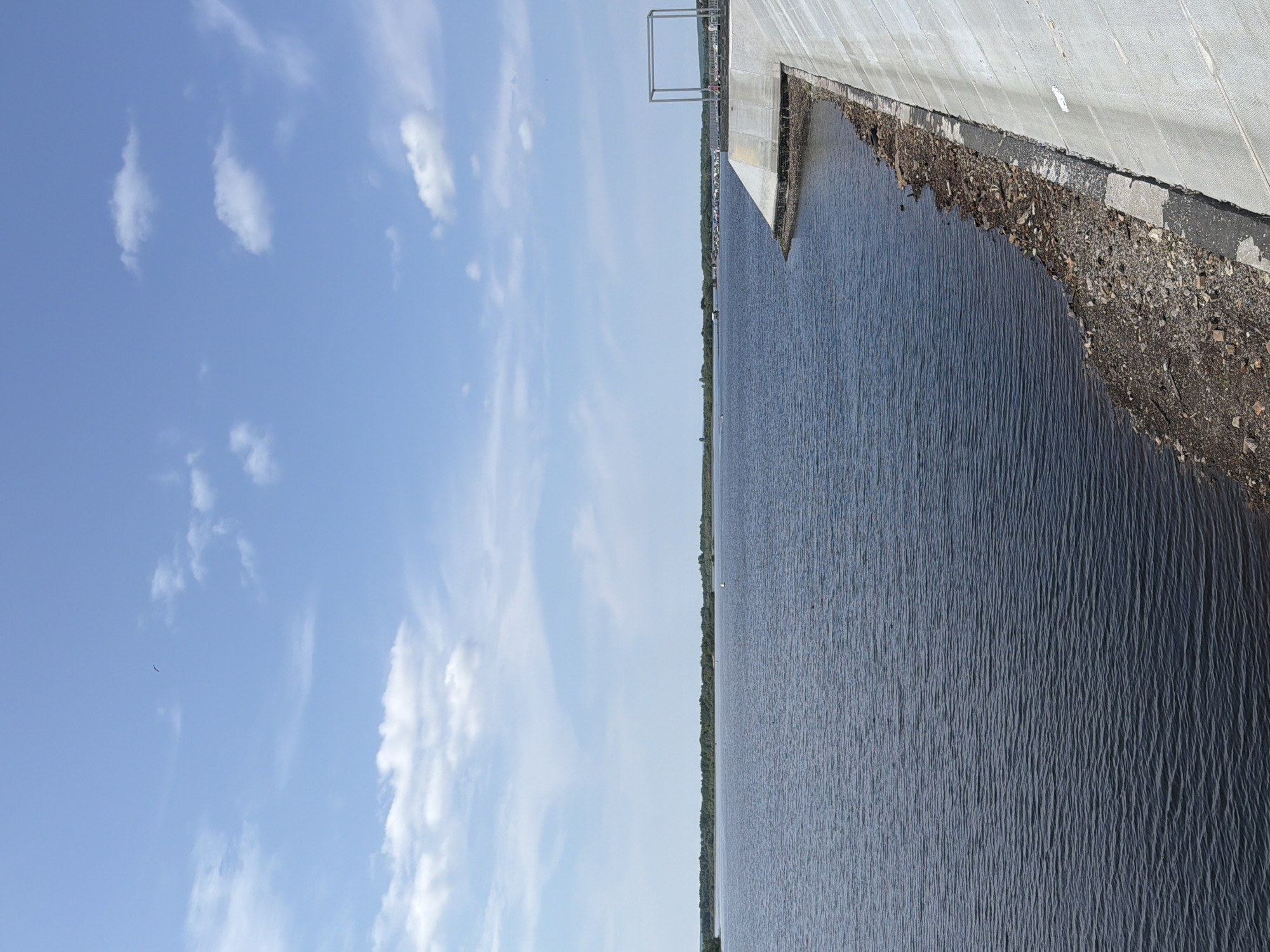 The image shows a body of water, likely a reservoir or a large river, under a blue sky with scattered white clouds. In the foreground, there is a concrete embankment sloping down to the water's edge, which is lined with rocks and debris. The water is dark blue with small ripples. In the distance, a treed shoreline is visible, and a few small boats or buoys are scattered on the water. On the embankment, a large, empty metal frame structure is visible. The scene appears to be a dam or a waterfront area. This location is identified as the Solikamsk reservoir in Solikamsk, Russia. There are no visible people in the frame.