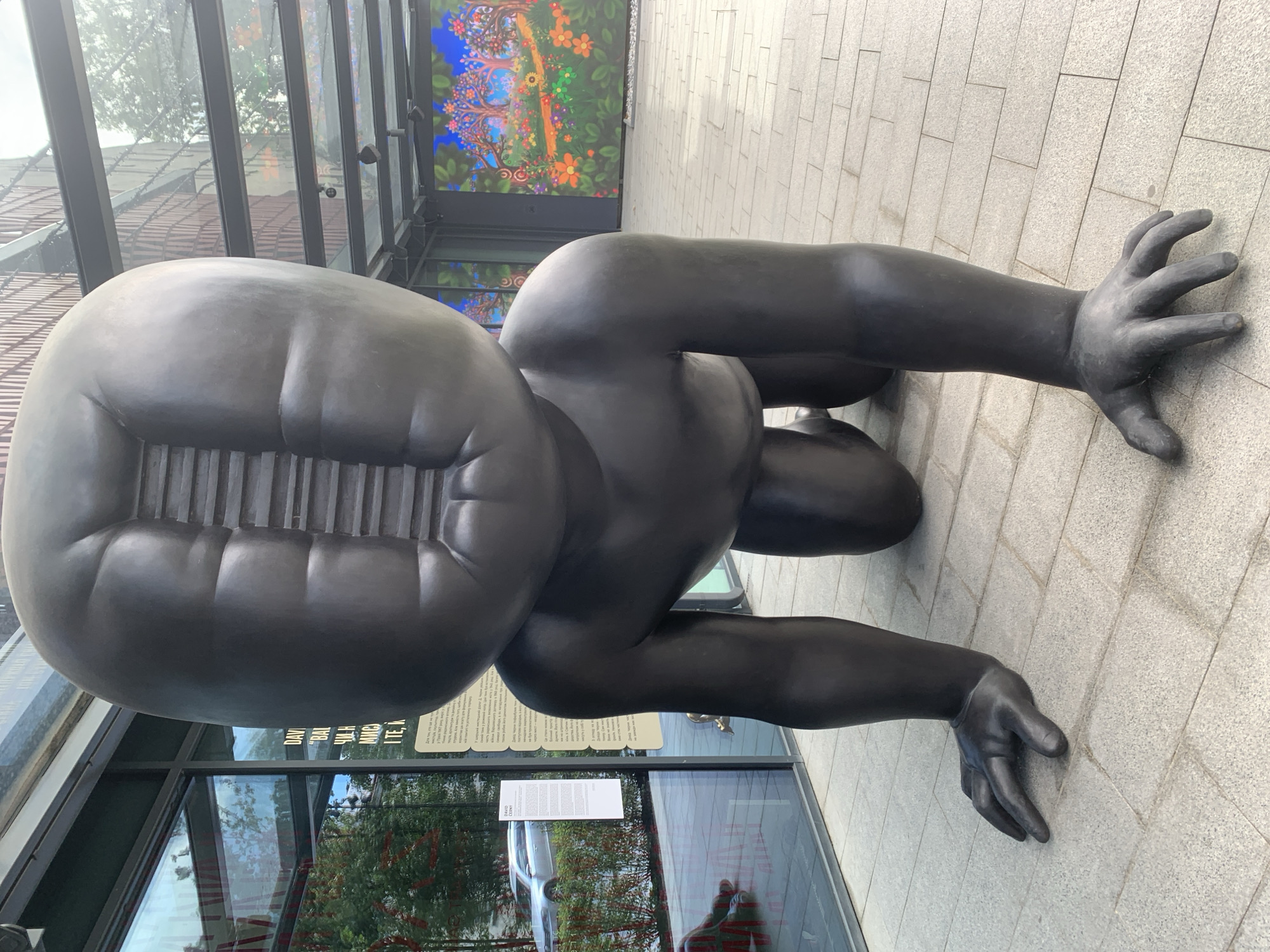 A large, dark bronze sculpture of a crawling baby with its head tilted back and a slit down the center of its forehead stands on a paved plaza in front of a modern building with large glass windows. The baby sculpture is on its hands and knees, with its fingers splayed on the ground. Reflected in the glass windows are trees and a silver car. To the right of the sculpture, a large, colorful digital mural displays a landscape with trees and flowers. A sign with text in Ukrainian is visible on a glass pane to the left of the sculpture. The plaza is paved with light-colored rectangular tiles.