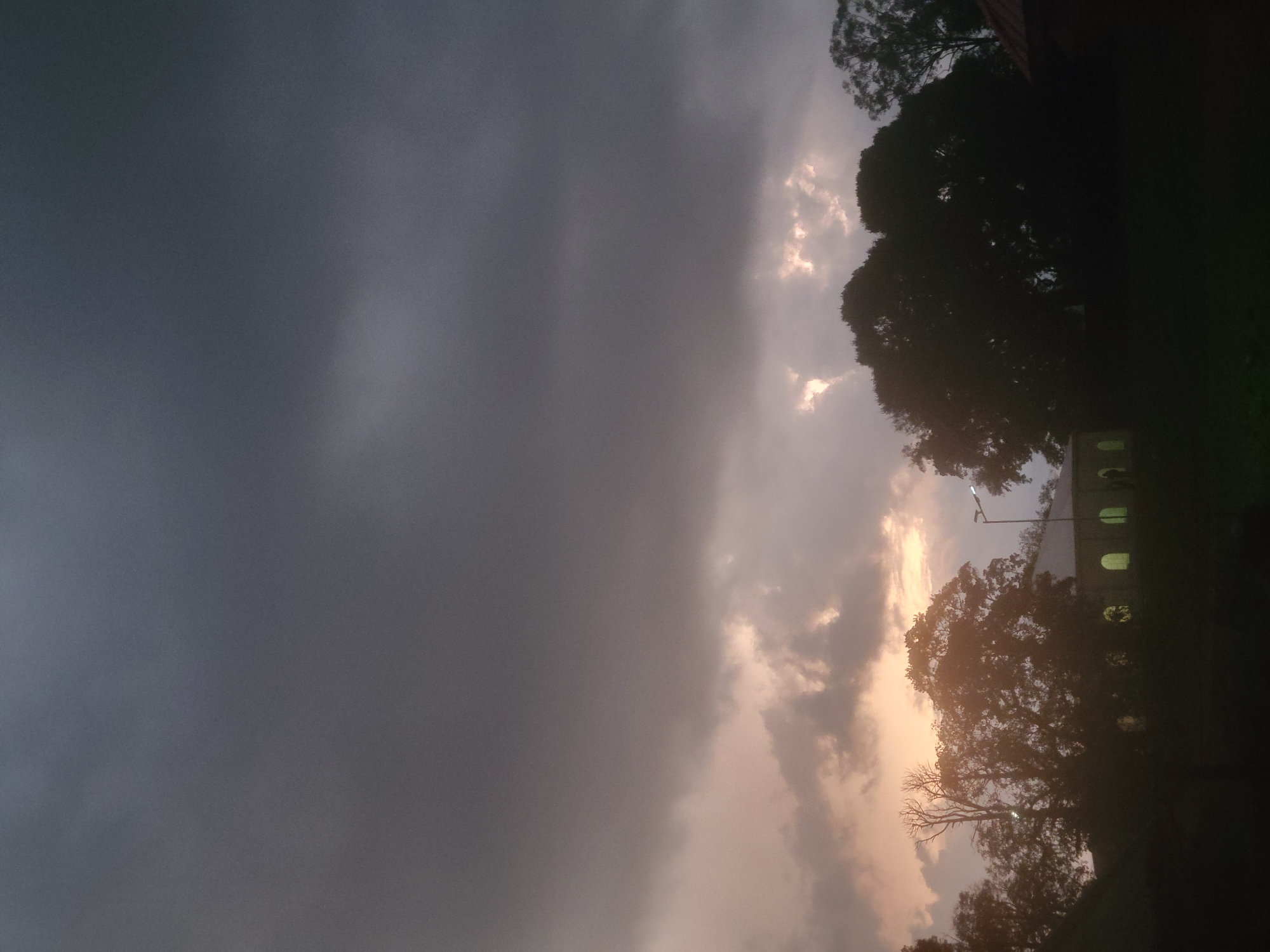 This image depicts an overcast twilight or dawn scene in Unknown, Unknown, dominated by a heavy cloud layer. The upper two-thirds of the frame consist of dark grey, uniform stratiform clouds. Towards the horizon in the lower third of the sky, the clouds are broken, revealing warm light, suggesting a low sun. This light casts hues of pale orange, pink, and yellow onto the cloud undersides, with several brighter, localized patches of light visible through the cloud breaks.
The lower third of the image is characterized by a dark, silhouetted landscape. A dense line of trees with varying foliage densities spans across the mid-ground. On the left side, a prominent tree with bare, gnarled branches stands out against the lighter sky. To the left of the center, a long, low building with a light-colored roof is partially visible, featuring a series of regularly spaced arched windows that emit a warm, interior artificial light. An illuminated street lamp, mounted on a pole, is positioned in the mid-ground between the trees and extends upwards into the lighter sky band. On the far right, a section of a reddish-brown tiled roof is partially visible. The immediate foreground is a dark, indistinct grassy area. No people or specific actions are discernible within the frame.