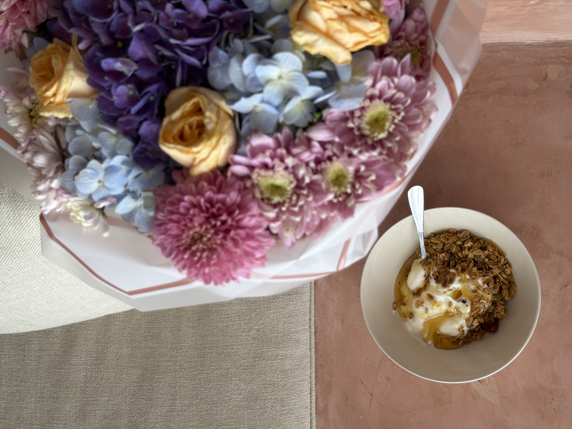 A high-angle shot reveals a scene indoors, combining a bouquet of flowers and a bowl of food. The bouquet on the left showcases roses, hydrangeas, and chrysanthemums in pastel shades. The bowl, positioned to the right, contains granola, yogurt, and honey, accompanied by a spoon. The surface beneath the bowl is light orange, while the bouquet rests on a cream-colored surface. It is likely taken in North Kuta, Indonesia.