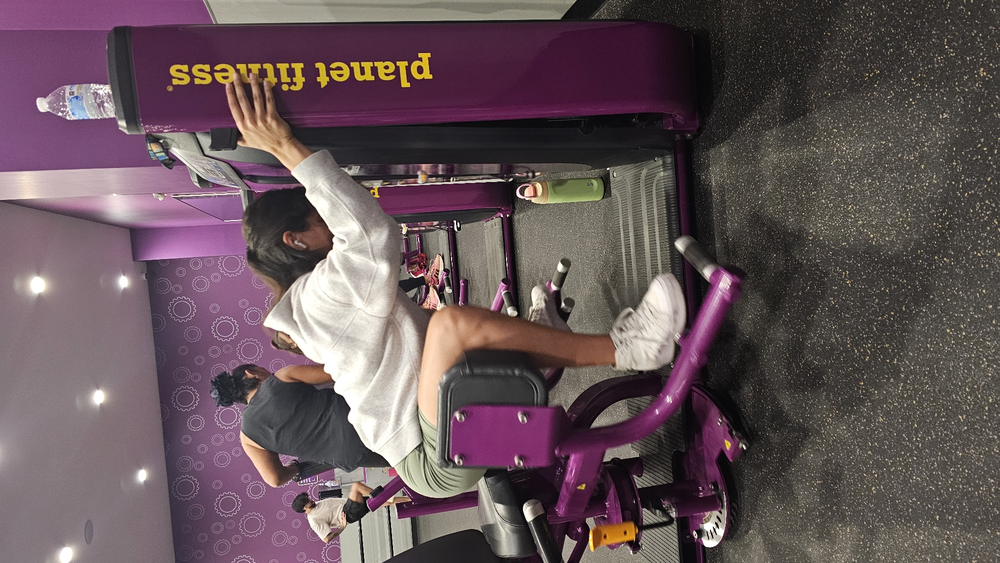The image depicts an interior view of a Planet Fitness gym, located in Puebla, Mexico. The foreground features an individual wearing a white hooded sweatshirt, olive green shorts, and white high-top athletic shoes, engaged in exercise. The person is bent forward, leaning with both hands on the vertical purple column of a gym machine, which prominently displays the yellow "Planet Fitness" logo. White wireless earbuds are visible in the person's ears. Their legs are positioned on the padded components of the purple and black exercise machine. A clear plastic water bottle rests on the top surface of the machine's column, and a light green reusable water bottle with a pink lid is on the speckled black rubber gym floor adjacent to the machine.
In the mid-ground, a second individual, wearing a black tank top with dark hair tied up, is partially visible from the back, standing near another purple exercise machine. Further in the background on the left, a third individual in a light-colored t-shirt and dark shorts stands with one hand on their hip. A fourth person's lower legs and pink athletic shoes are partially visible in the right background.
The gym environment includes purple walls; the left wall features a pattern of white interconnected gear-like circles, while the right wall is solid. The white ceiling contains several recessed circular lights.