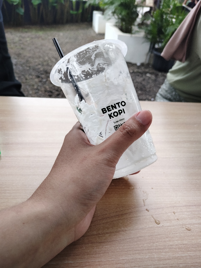 The image displays a close-up of a person's hand holding a clear plastic cup with a black straw, set against a blurred background. The cup, which is mostly empty, shows white residue from a consumed beverage and contains melting ice. On its side, the brand "BENTO KOPI" is prominently printed, along with the text "Scan Here" and a QR code. Below the cup, on the light-colored wooden table, are several small droplets of liquid, likely condensation or a spill.
In the out-of-focus background, an outdoor or semi-outdoor cafe setting is visible, featuring green plants in white planters and on the ground, which appears to be gravel or concrete. A blurry figure of another person, wearing a green top and a light-colored headscarf, is partially seen on the right. The scene depicts a casual moment during the daytime at a cafe or similar establishment in Dramaga, Indonesia, following the consumption of a cold drink.