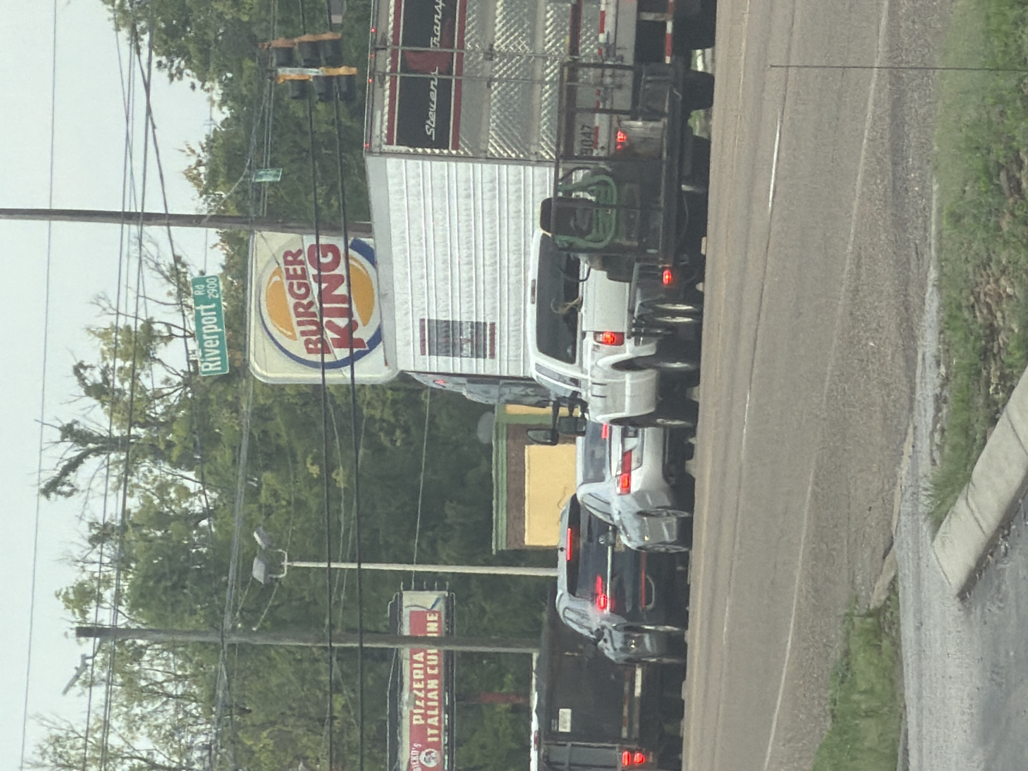 The image shows a street scene with several vehicles in traffic. In the background, there is a Burger King restaurant and a sign for "Riverport Rd 2900". There is also a sign for "Dickey's Pizzeria Italian Cuisine". In the foreground, there are several cars and trucks, including a white pickup truck pulling a trailer with a green piece of equipment on it. The sky is overcast. The scene appears to be in an urban or suburban area. The Burger King sign is a strong indicator that this is likely in a commercial district. The presence of traffic lights and multiple lanes of traffic suggest a busy intersection. The overall impression is a snapshot of everyday life on a road in a city.