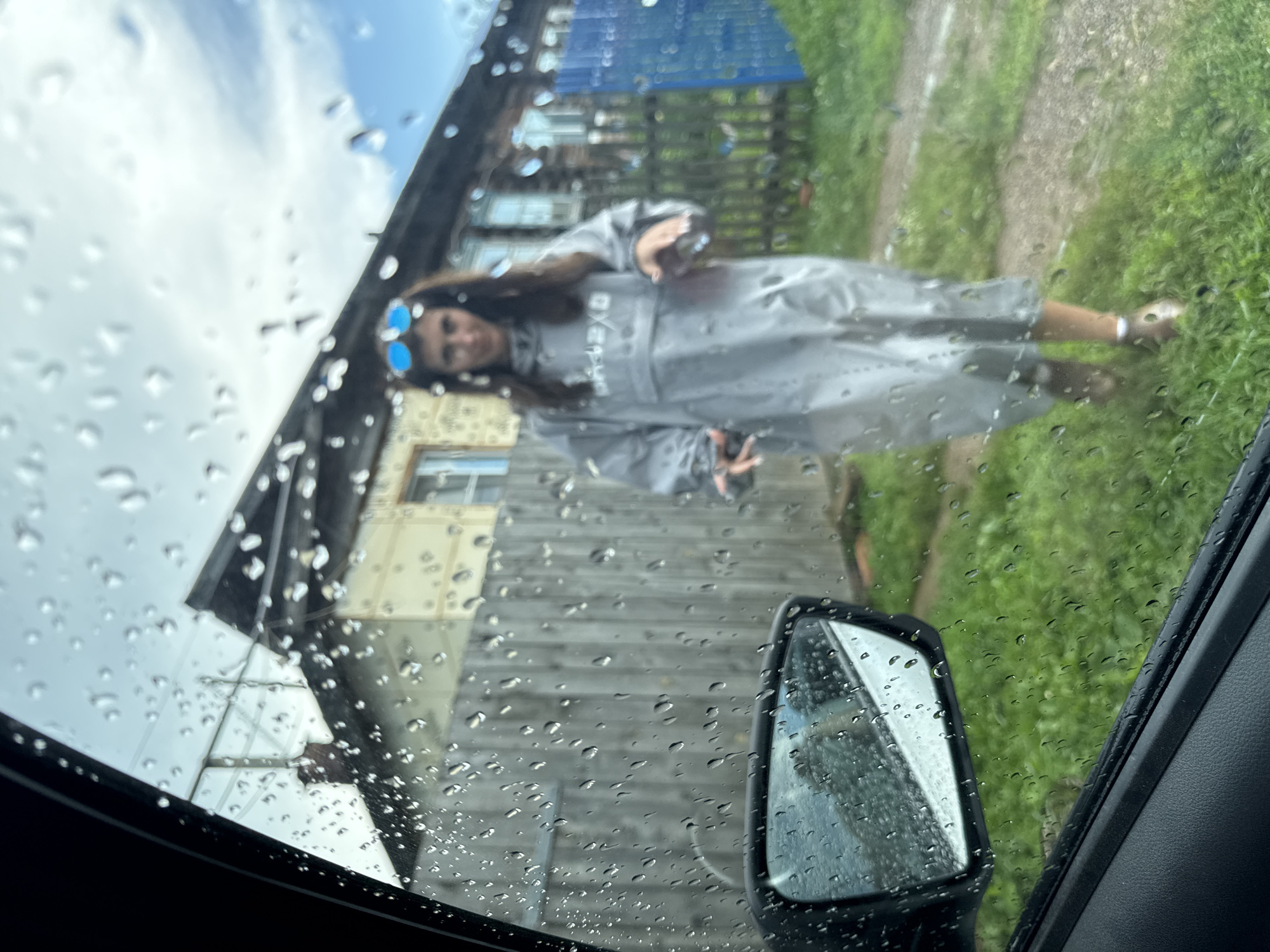 This is a medium shot taken from inside a vehicle, looking out through a rain-streaked windshield. In the foreground, the black interior of the car and a portion of the side mirror are visible. The side mirror reflects a blurred image of green grass and the sky.
The background depicts a rural scene, likely in the city of Vasya, Russia, with a wooden fence and a two-story building that appears to be a traditional wooden house. The house has multiple windows and a chimney. A bright blue fence is visible to the right of the house.
In the middle ground, a young woman is walking towards the camera. She is wearing a long, light grey poncho or raincoat and blue sunglasses. Her hands are visible, and she appears to be holding something in her right hand. The ground is covered in green grass and a dirt path. The overall atmosphere suggests a cloudy or rainy day.