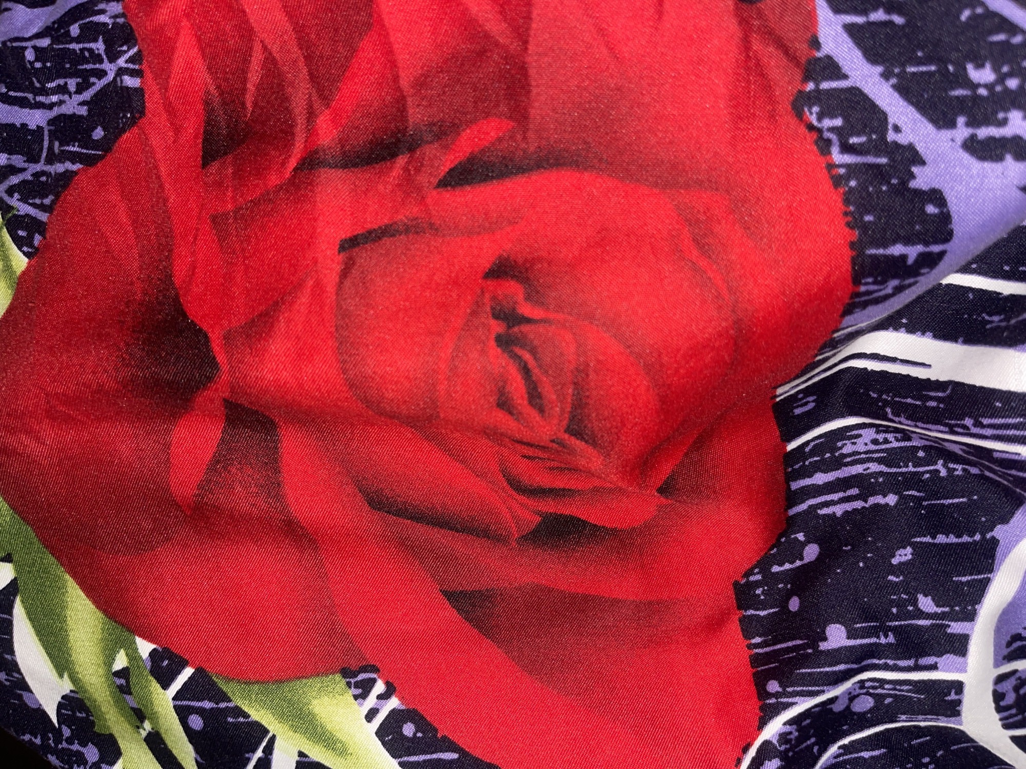 The image is a close-up, high-angle photograph of a fabric with a floral pattern. The dominant element is a large, abstract red rose, rendered in a soft, painterly style. The rose is partially unfurled, with its petals creating layers of vibrant red and deep shadows. The background of the fabric features a dark, abstract pattern with streaks and splatters of purple and white against a black base. To the left of the rose, portions of green leaves are visible, also in a stylized, artistic rendering. The texture of the fabric appears to be a smooth, possibly synthetic material, with a subtle sheen that highlights the folds and creases. The lighting is direct, creating strong contrasts and emphasizing the textural details of the fabric.
There is no visible indication of the city of Minsk, Belarus, in this image, nor are there any people, actions, interactions, or environmental elements that would suggest a specific location within a city. The photograph is solely focused on the detailed pattern of the fabric itself.