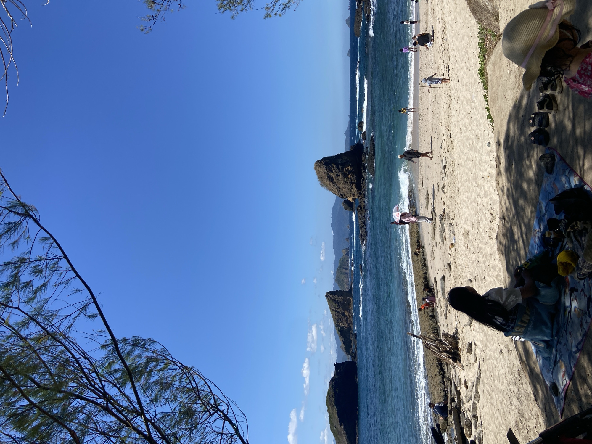 The image depicts a coastal scene at a beach. The location is in Unknown, Indonesia. A sandy beach is occupied by several people. Some are walking along the shoreline, while one is seated on a patterned blanket. Visible near the seated person are footwear and a hat. The beach meets the ocean, which has small waves. Rock formations are visible within the ocean. The sky is a clear blue. Foliage is present in the foreground.
