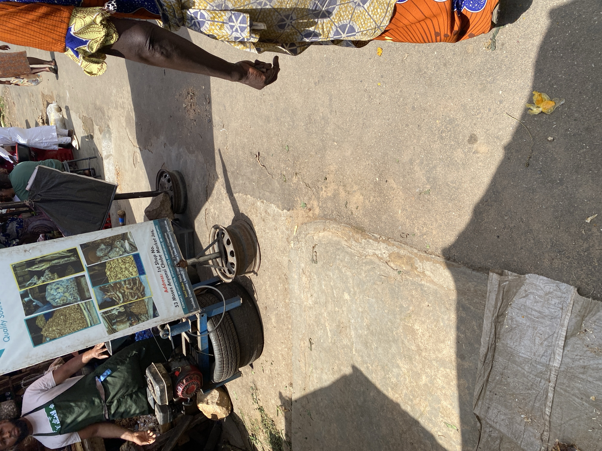 The image shows a street scene in Jos, Nigeria, likely a market area. In the foreground, a man wearing a white t-shirt and a green apron stands next to a small engine. Behind him, a large sign displays images of food items and text, including an address for "1st Shop No. 53 Moses Anyansi Chobe Market Jos," and contact information. To the right, a person in a vibrant orange and blue patterned dress is partially visible, walking away from the camera. The ground is paved with concrete and asphalt, with some debris and shadows present. Several tires are stacked near the engine. In the background, other individuals and market stalls can be seen, suggesting a busy commercial environment.