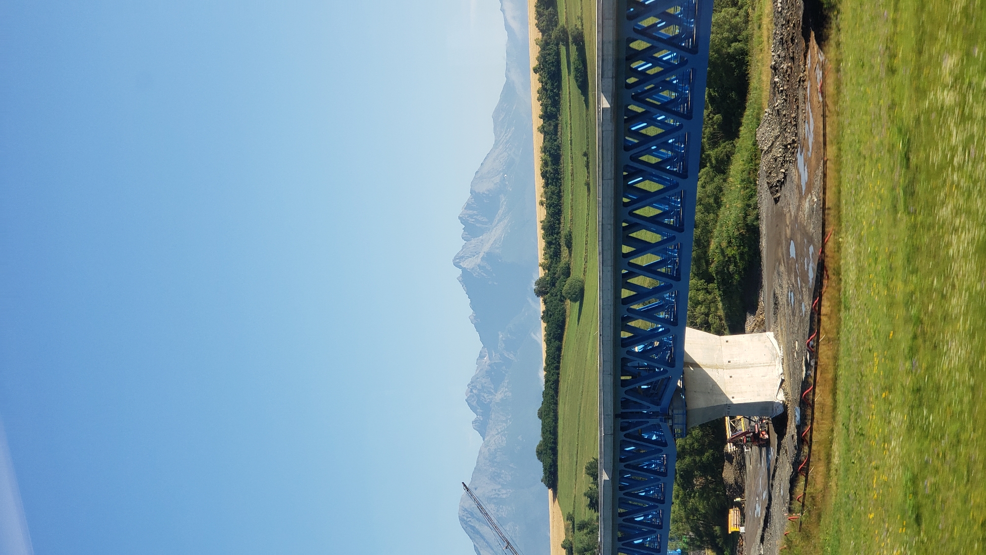 The image captures a scenic view of the High Tatras mountains in the background, with a bright blue sky above. In the foreground, a large blue truss bridge under construction is visible, supported by a concrete pillar. The bridge spans across a landscape that includes green fields, trees, and a body of water. The area around the bridge appears to be a construction site, with piles of gravel, construction vehicles, and what looks like red piping on the ground. In the mid-ground, there are more fields and trees, with the majestic mountain range forming a dramatic backdrop. The overall impression is one of natural beauty juxtaposed with ongoing infrastructure development in the vicinity of Gánovce, Slovakia.