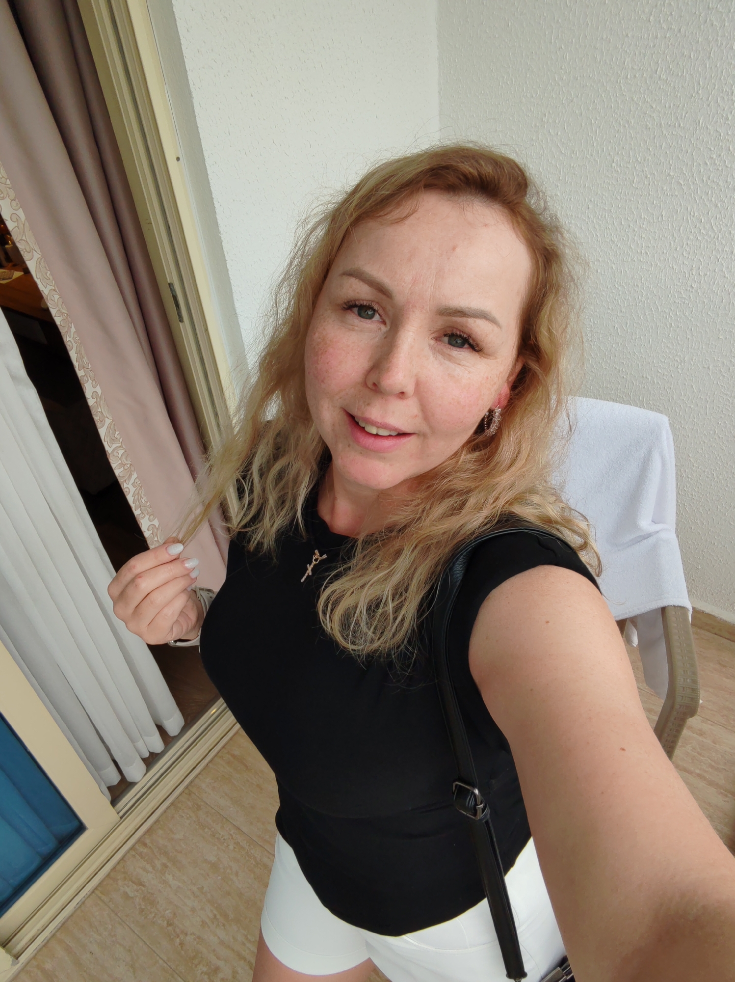 The image is a selfie taken indoors, likely in a hotel room or apartment, in Alanya, Turkey. A woman with blonde hair, freckles, and fair skin is the primary subject. She is wearing a black top and white shorts, and a black bag is slung over her shoulder. The background includes a window or sliding glass door with white and beige curtains, and a balcony wall with a textured surface. A chair with a white towel draped over it is also visible. The floor appears to be tiled. The woman is holding a strand of her hair in her left hand. She is looking directly at the camera with a slight smile.