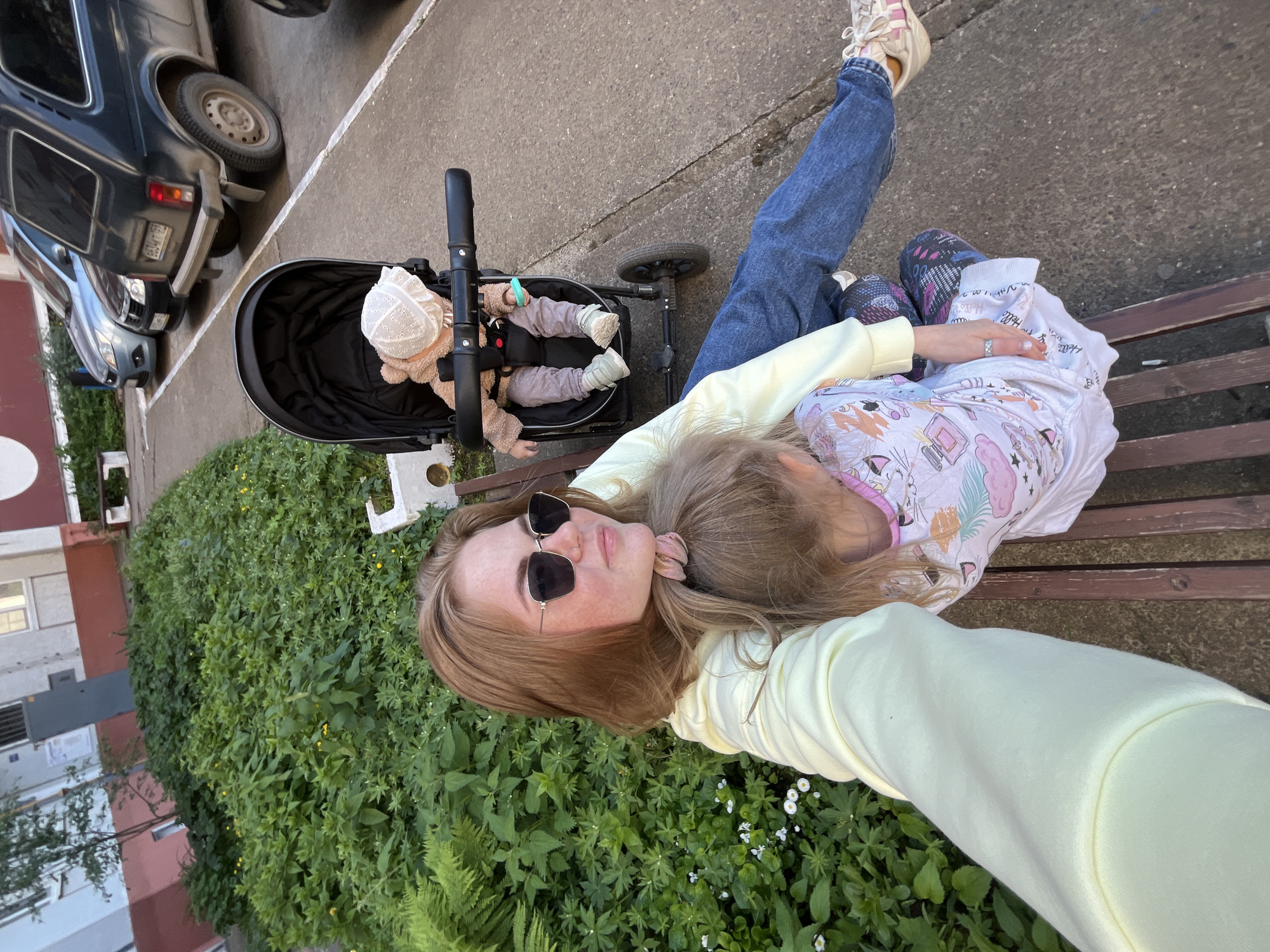 A woman with red-blonde hair and wearing sunglasses is lying on a wooden bench with a child resting on her, in what appears to be a selfie. The woman wears a light yellow long-sleeved shirt and blue jeans. The child is wearing a printed white shirt and patterned leggings. Beside the bench is a baby stroller with a baby inside wearing a white bonnet and brown outfit. The location features a building on the left and lush green plants in the foreground. There are cars parked nearby. This picture was taken in Chaikovsky, Russia.