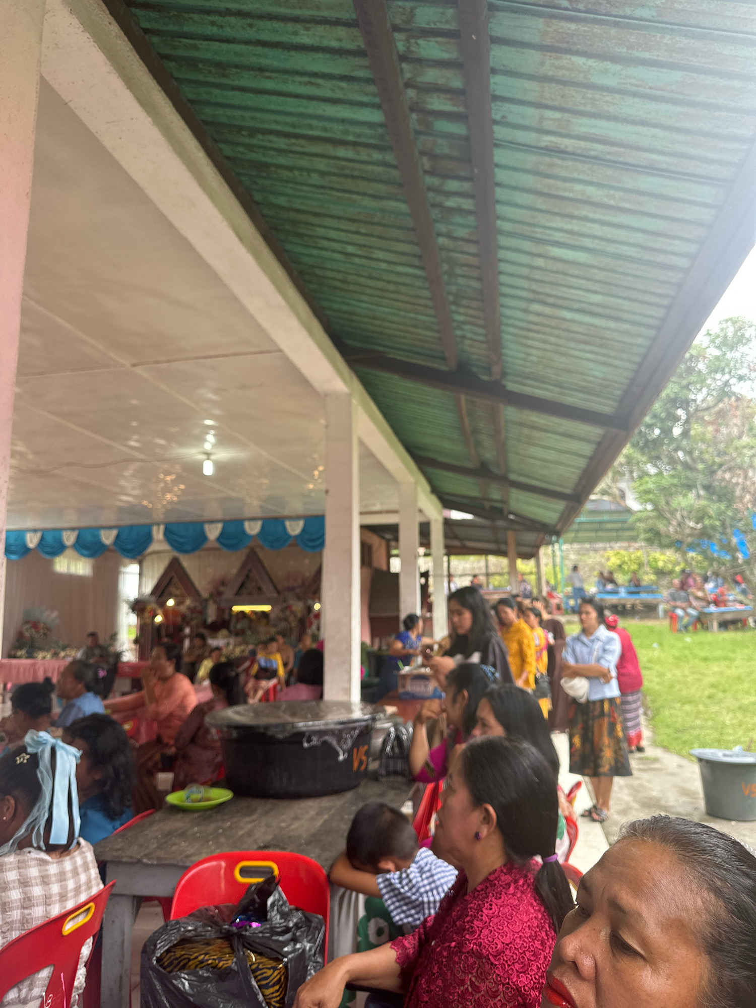 In the city of Pardamean Ajibata, Indonesia, an outdoor gathering is taking place under a green corrugated metal roof supported by white pillars. The scene depicts several individuals seated at a table in the foreground, with a focus on a woman wearing a pink patterned shirt and another wearing a red lace top. A large black basin or pot sits on the table, and next to it is a green dish. Red plastic chairs are visible, one with a black plastic bag containing what appears to be fabric or clothing. In the background, more people are seated, and a decorated stage or altar area with blue and white drapes is visible. Beyond this, a grassy area with trees and further seating suggests a larger event. The lighting appears to be natural, with some artificial lights visible within the covered area.