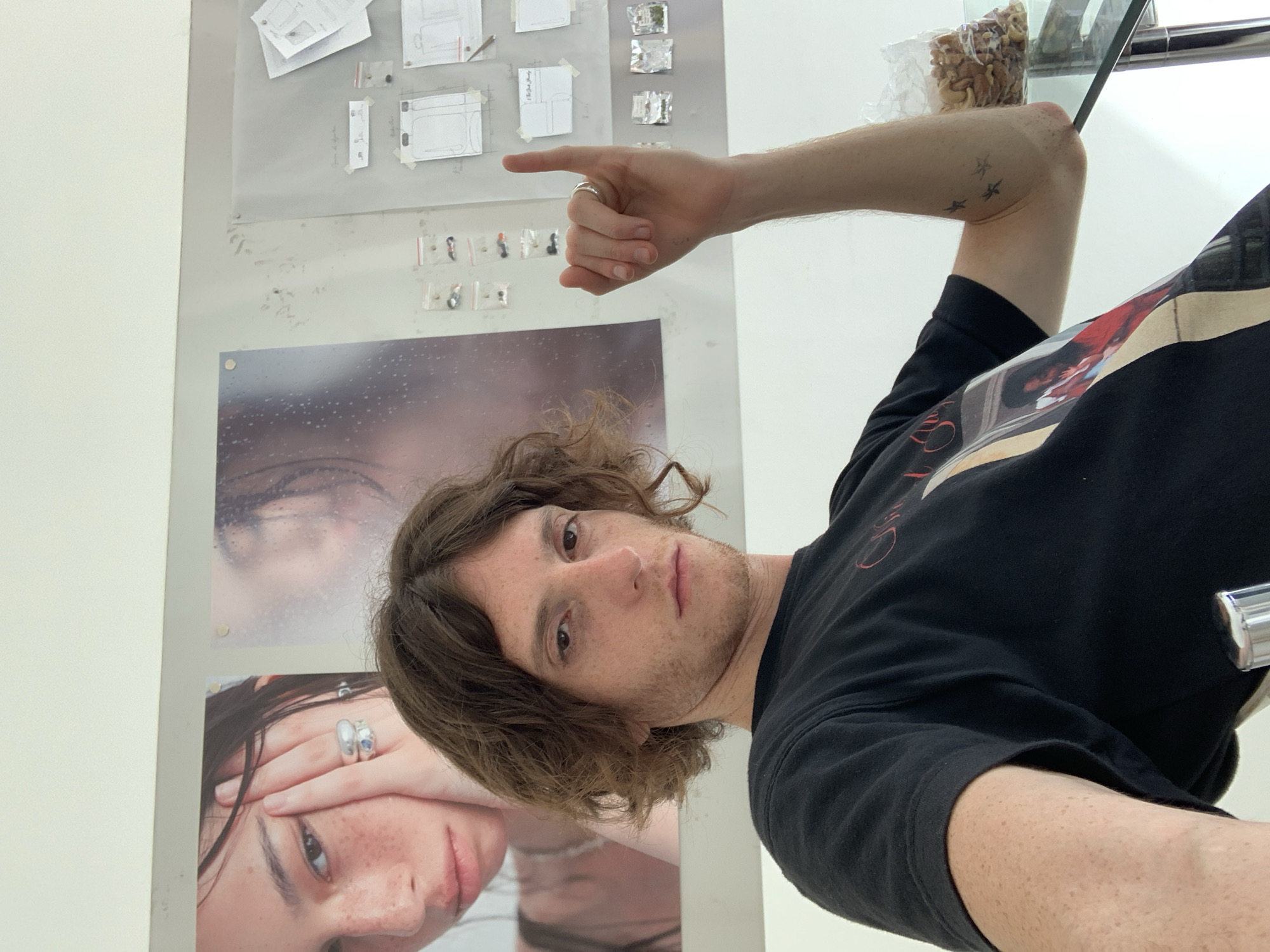A man with brown hair and a black t-shirt is pointing his finger towards a wall with taped up papers and photographs. The man has a ring on his finger and a tattoo of three stars on his arm. A glass shelf with a container of nuts is visible in the upper right corner. The location is not discernible from the image.
