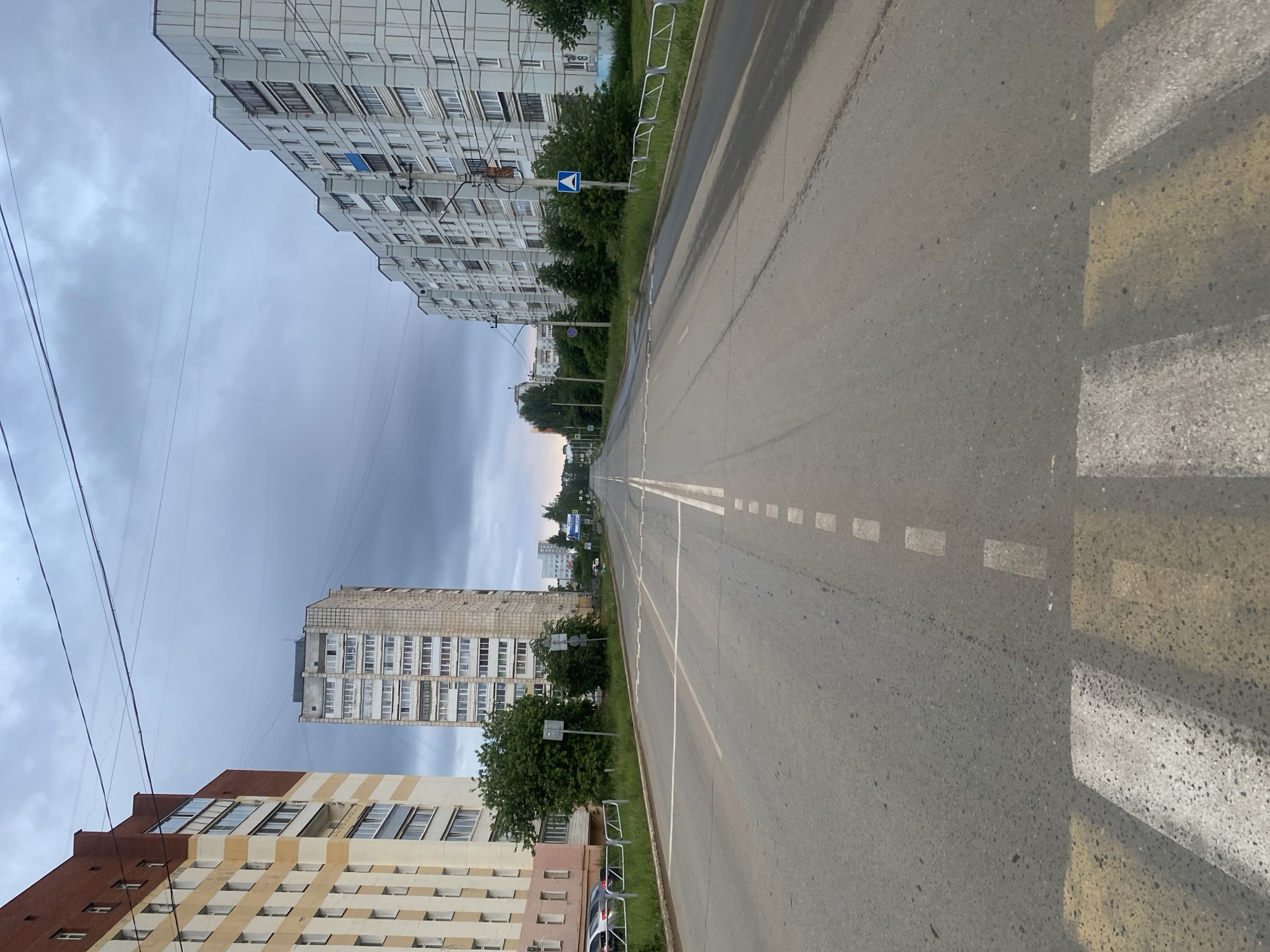 This street-level image, oriented vertically, captures a long, straight asphalt road lined by multi-story residential buildings under an overcast sky in Chaikovsky, Russia.
The road, appearing empty of traffic, features white dashed lines marking lanes and a solid white line along its edge. In the foreground on the right, a pedestrian crossing is clearly visible with its characteristic yellow and white striped markings.
On the right side of the street, a tall, light grey apartment building with numerous windows and balconies dominates the view. Closer to the road, a strip of green grass, bushes, and trees separates the sidewalk area from the street, along with silver metal safety railings. A blue and white road sign, displaying a white arrow pointing forward within a blue circle, is mounted on a pole amidst the greenery.
On the left side of the street, another multi-story building with a facade of brown and beige horizontal stripes is visible in the foreground. Further down the street on the left, a taller, lighter-colored apartment building stands. Trees and bushes also line this side of the road.
Overhead, multiple power lines crisscross against the grey, cloudy sky. The lighting suggests either early morning or late afternoon, as there are no harsh shadows, and a subtle brighter band is visible on the horizon. The street is devoid of people or vehicles, conveying a sense of quiet and stillness. No legible text is visible in the image.