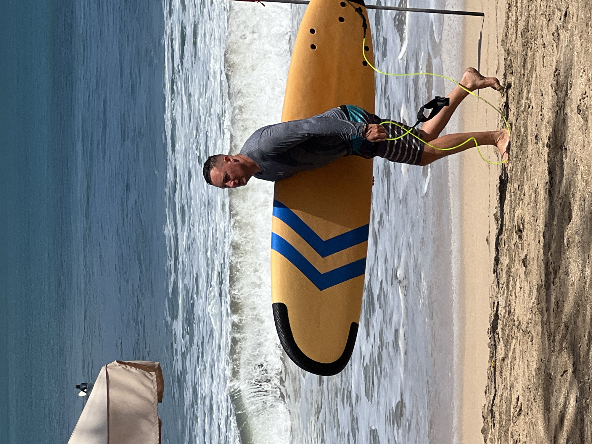 A man walks on a sandy beach in Kuta, Indonesia, carrying a yellow surfboard with blue accents. He is wearing a grey long-sleeved shirt and striped shorts. The surfboard has a yellow leash attached to it, which he is holding. Waves are breaking in the background, and a beige umbrella is visible to the left. The sky is clear and blue.