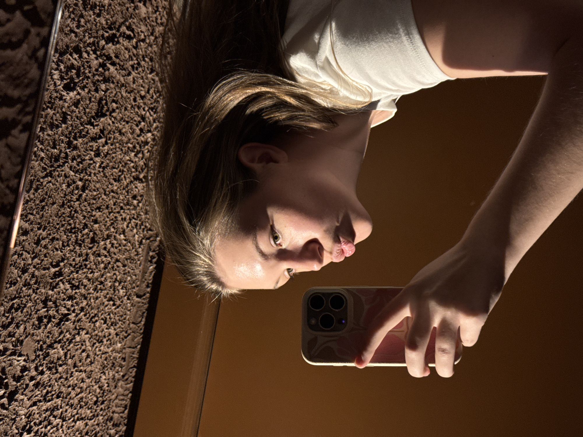 A selfie of a young woman is reflected in a mirror. The woman is holding an iPhone, its triple camera array visible. She is pouting her lips. Her hair is a mix of blonde and light brown. She's wearing a white shirt. The background includes a textured, dark-colored wall or doorframe. The reflected environment appears to be a room with an orange-brown wall. It is not possible to determine if the location is Belgrade, Serbia, from the given image alone.